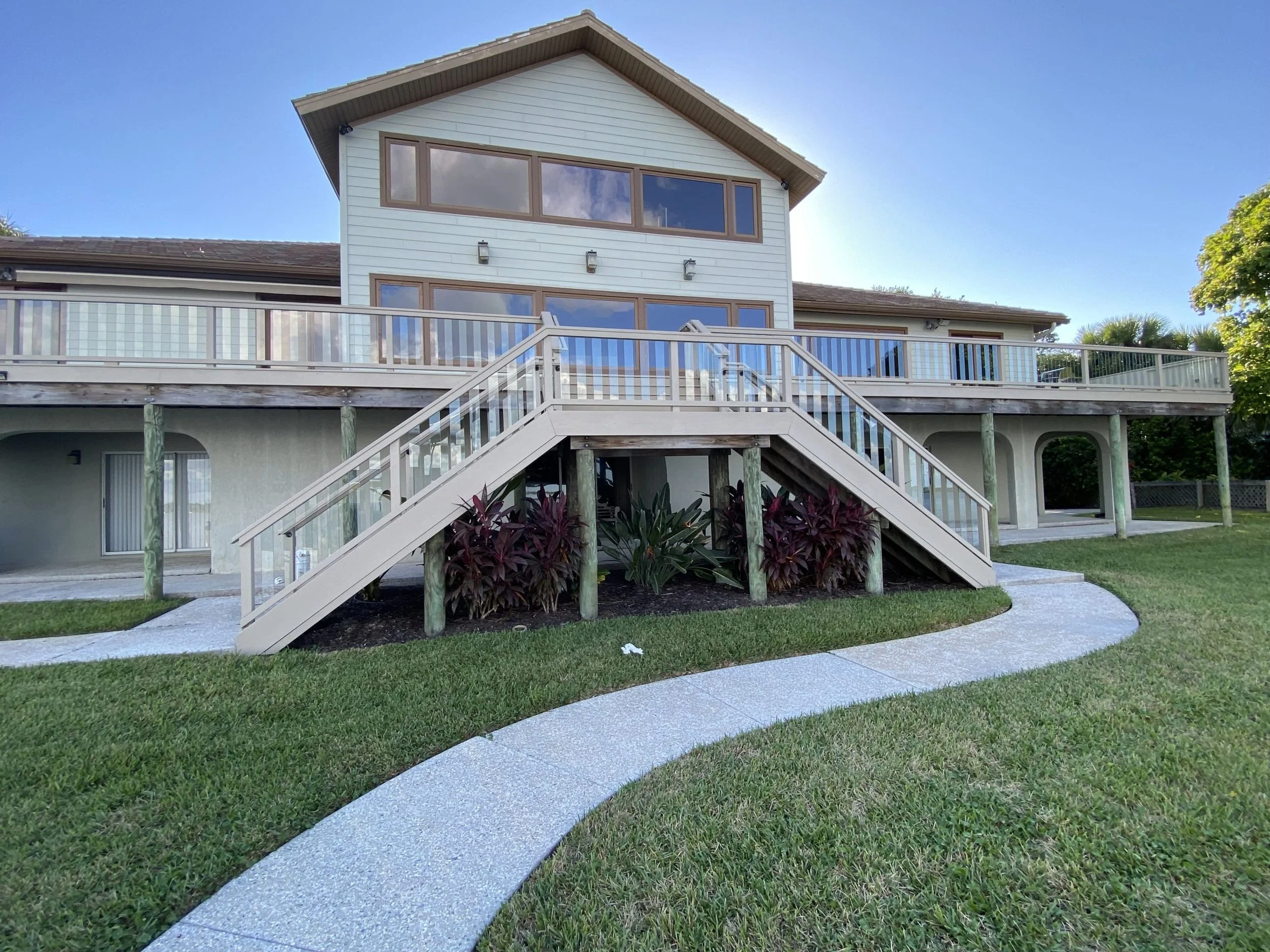Back view of a two-story house with a staircase leading to a balcony, surrounded by green lawn and trees.