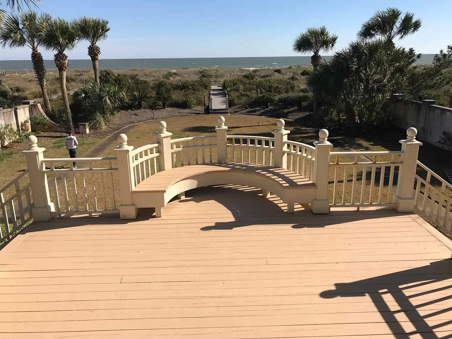 View of a wooden deck with a curved white railing, overlooking a grassy outdoor area with palm trees and a pathway leading to the beach in the background.