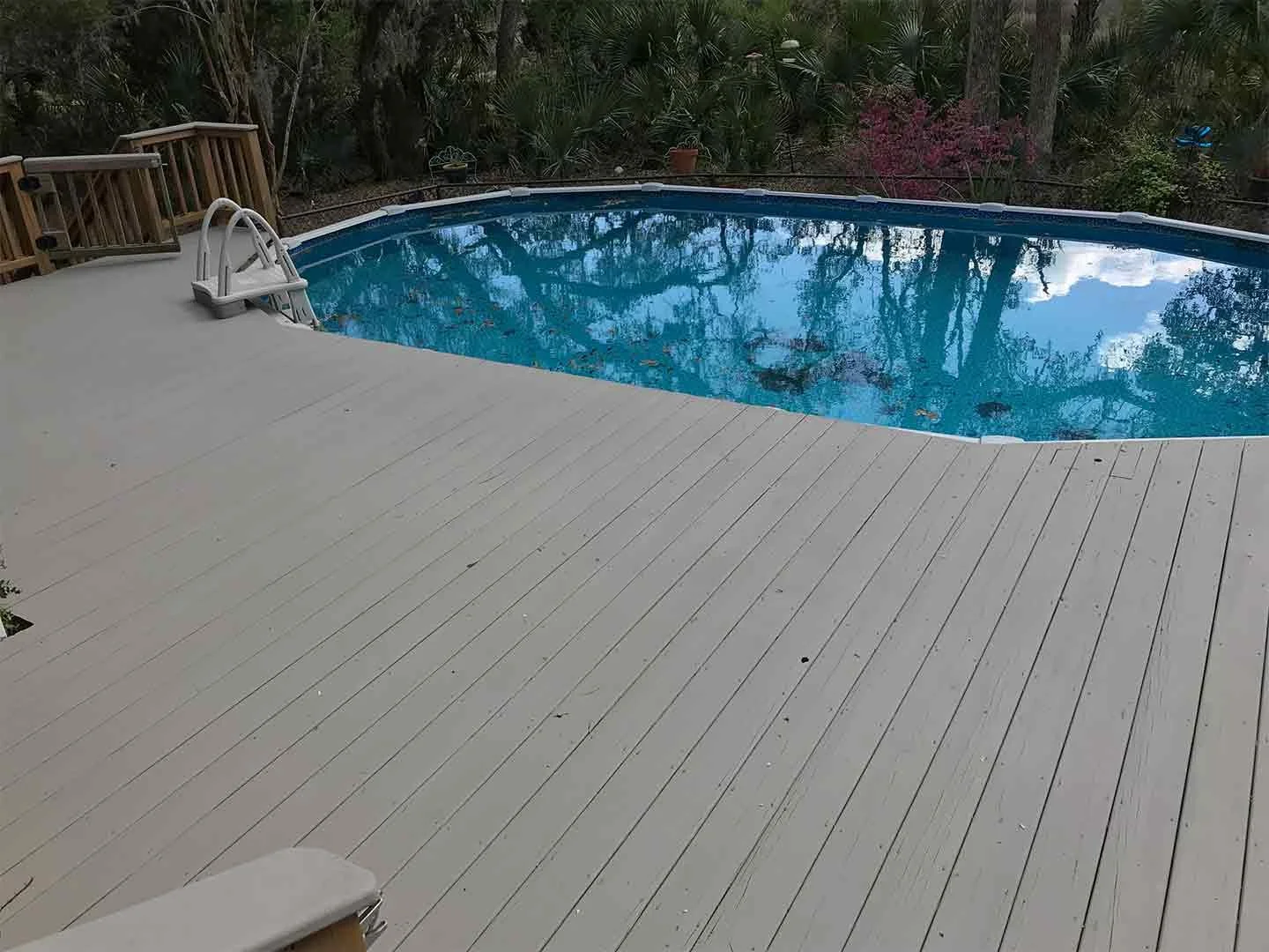 An outdoor backyard scene featuring a swimming pool with a white wooden deck, a metal ladder at the pool's edge, and a backdrop of trees and shrubs.