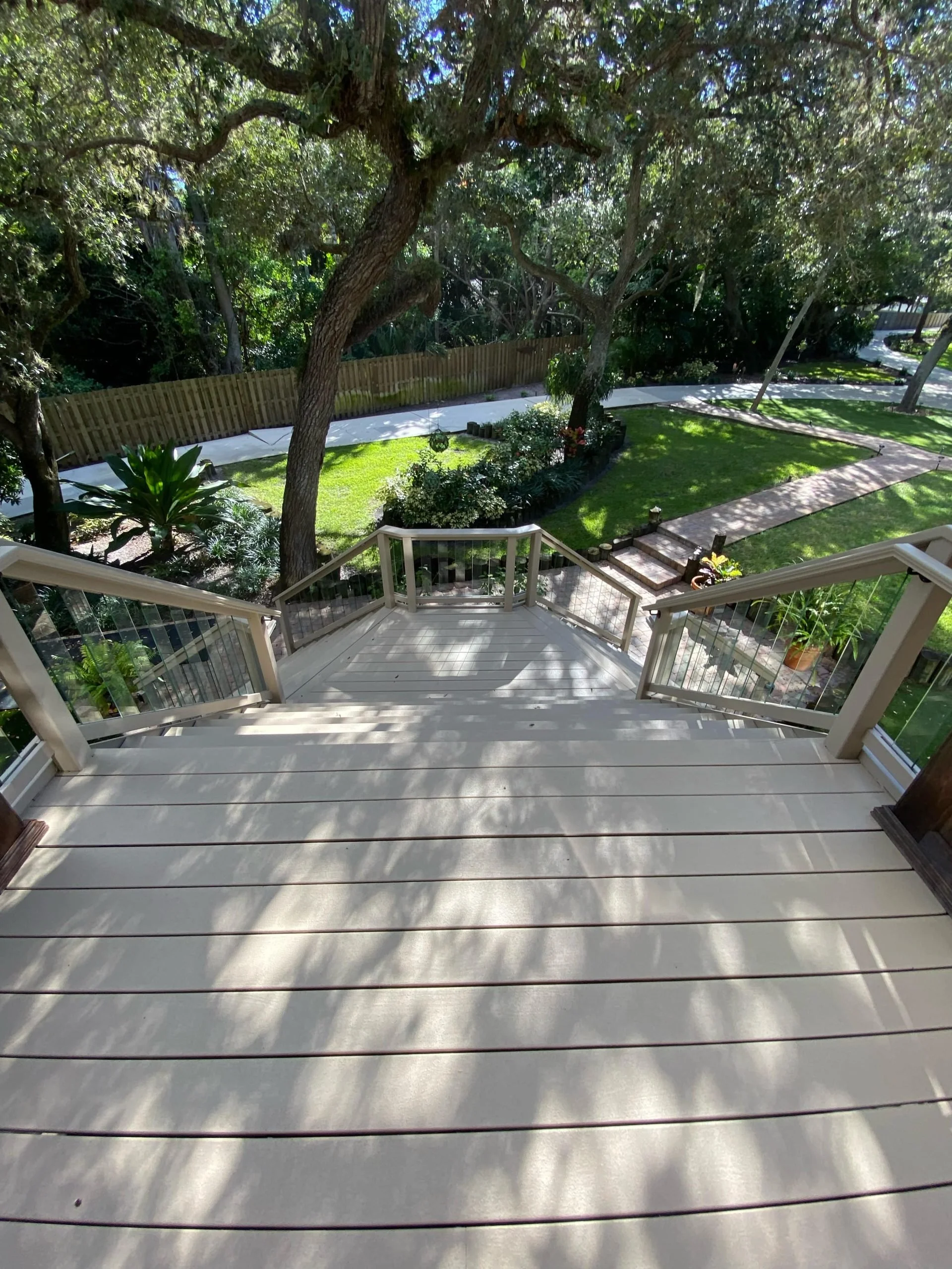 View from a porch staircase overlooking a backyard garden with green grass, trees, flowering bushes, a curved dirt pathway, and a wooden fence.