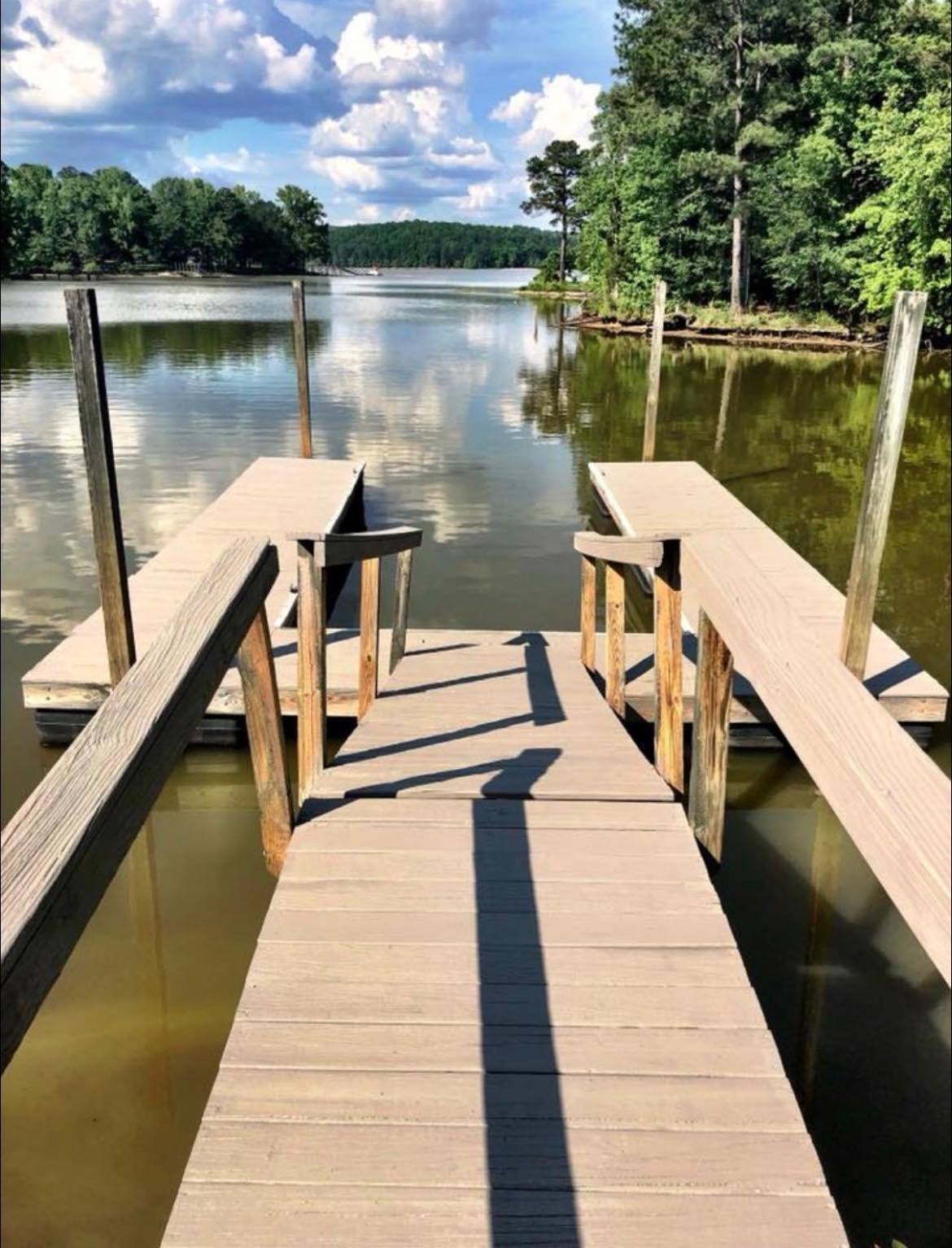 A wooden dock extends into a calm lake with surrounding trees and a partly cloudy sky.