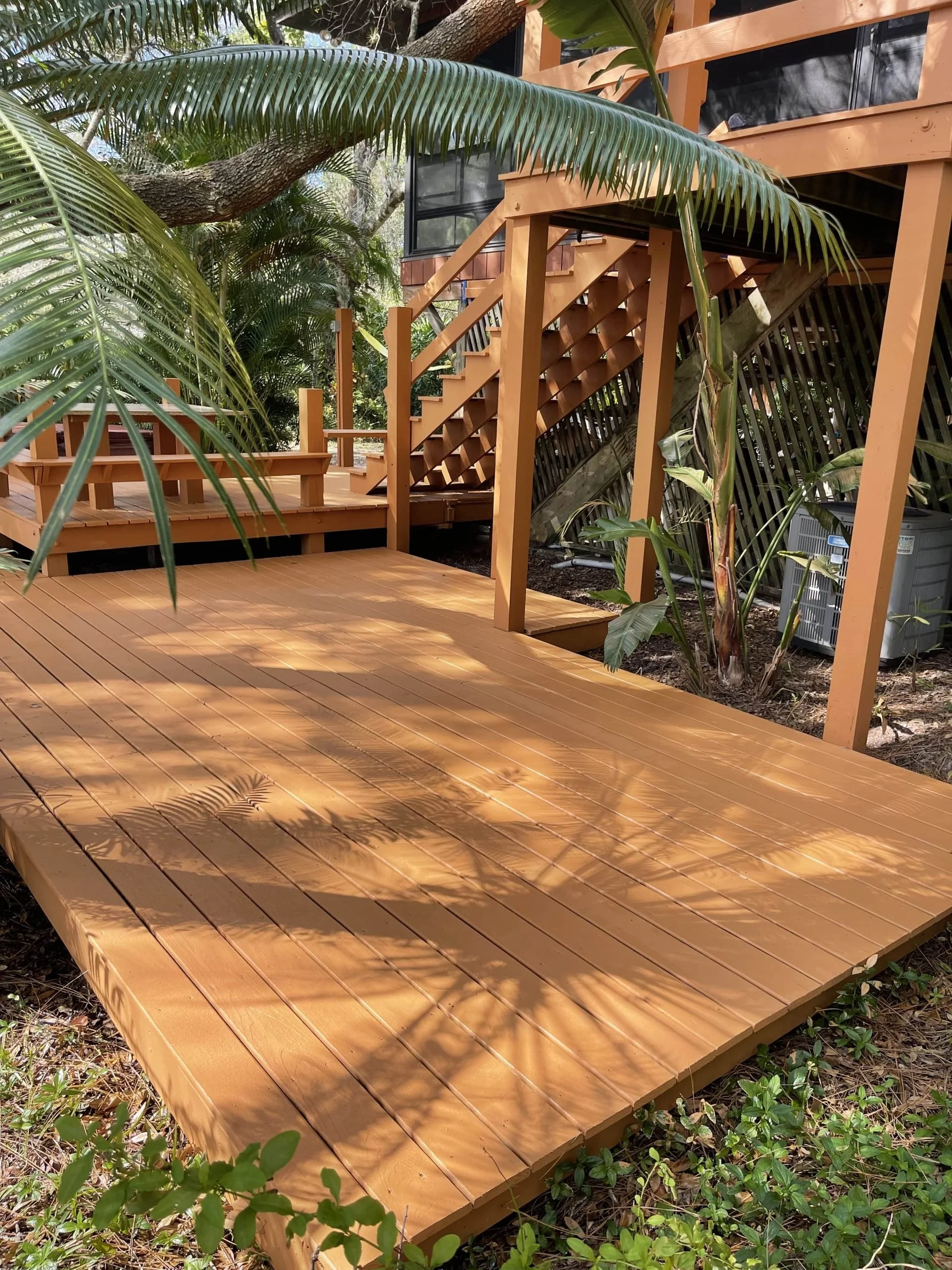 Wooden deck with stairs leading to an elevated area, surrounded by tropical plants and trees.