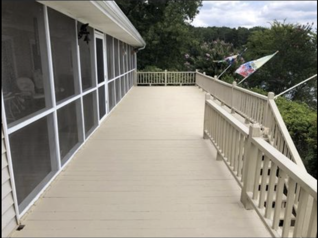 A spacious porch with beige wooden flooring and white railing, attached to a house with large screened windows on the left. In the background, there are trees and a cloudy sky, with two colorful umbrellas visible beyond the railing.