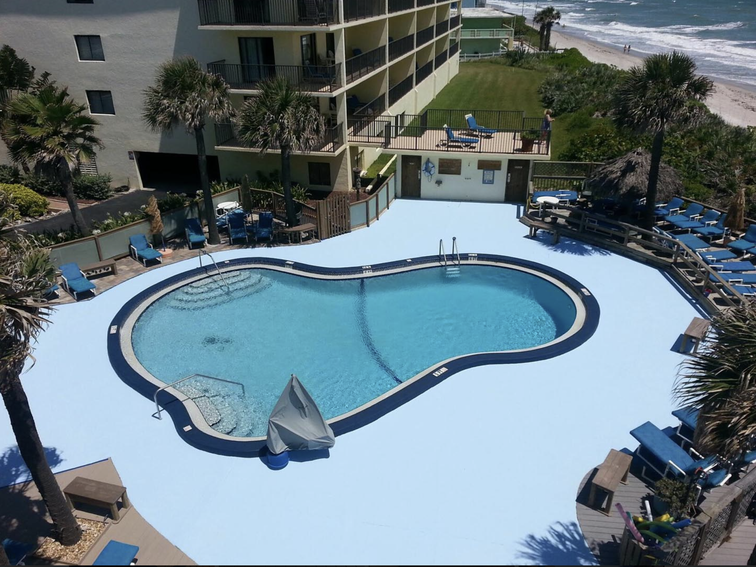 An aerial view of a swimming pool surrounded by lounge chairs and trees near a beach with sand and ocean waves. A building with balconies overlooks the pool area.