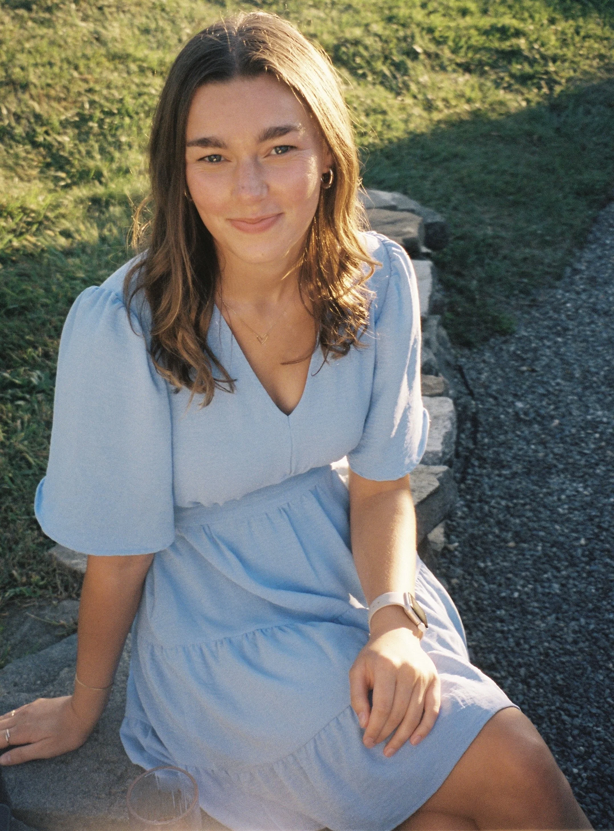 A young woman with shoulder-length brown hair, wearing glasses, a brown top with ruffled sleeves, and a gold necklace, smiling at the camera against a white background.