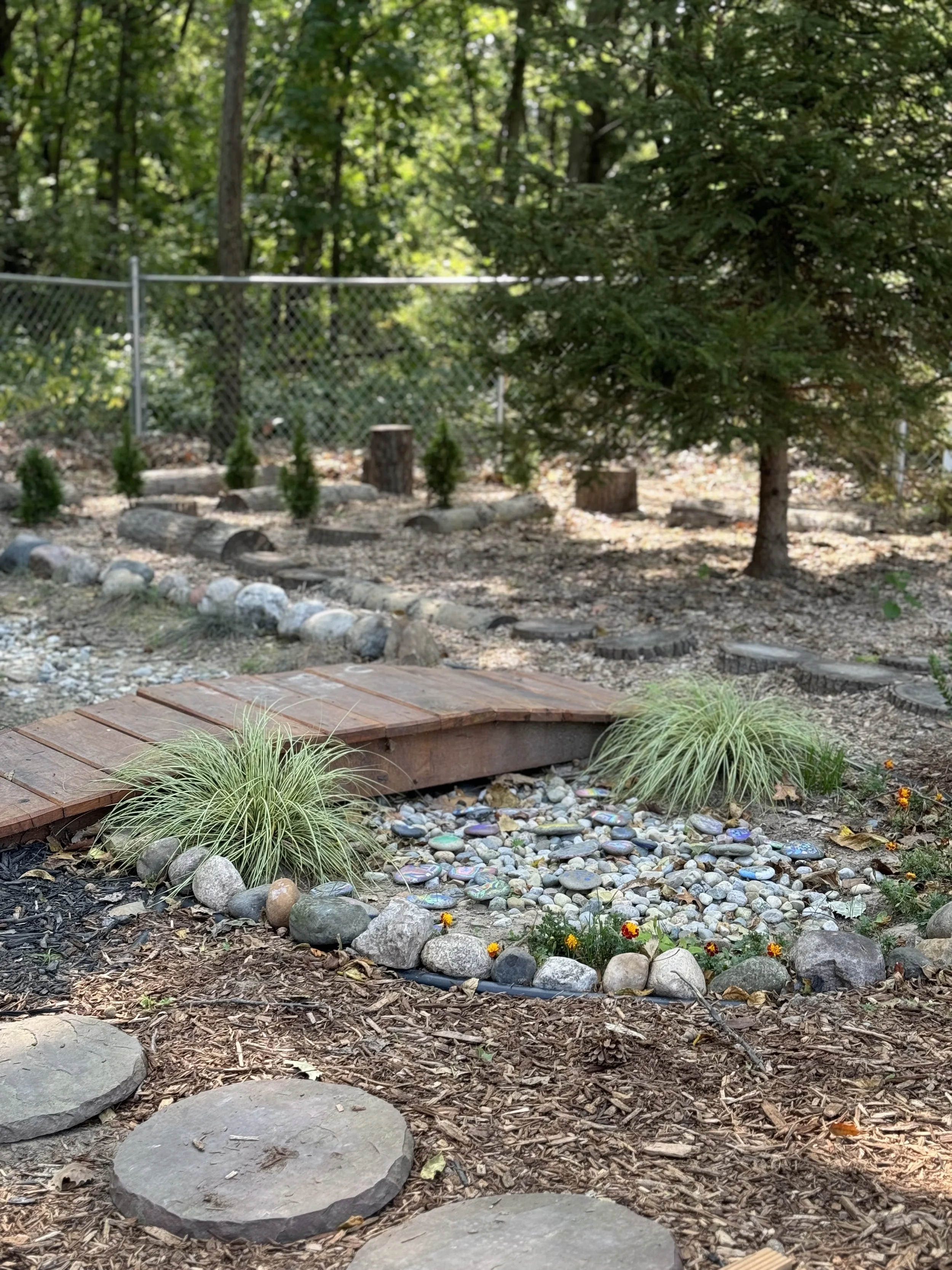 A small garden area with a wooden bridge over a dry decorative creek bed filled with colorful painted rocks, surrounded by plants, trees, and a chain-link fence in the background.