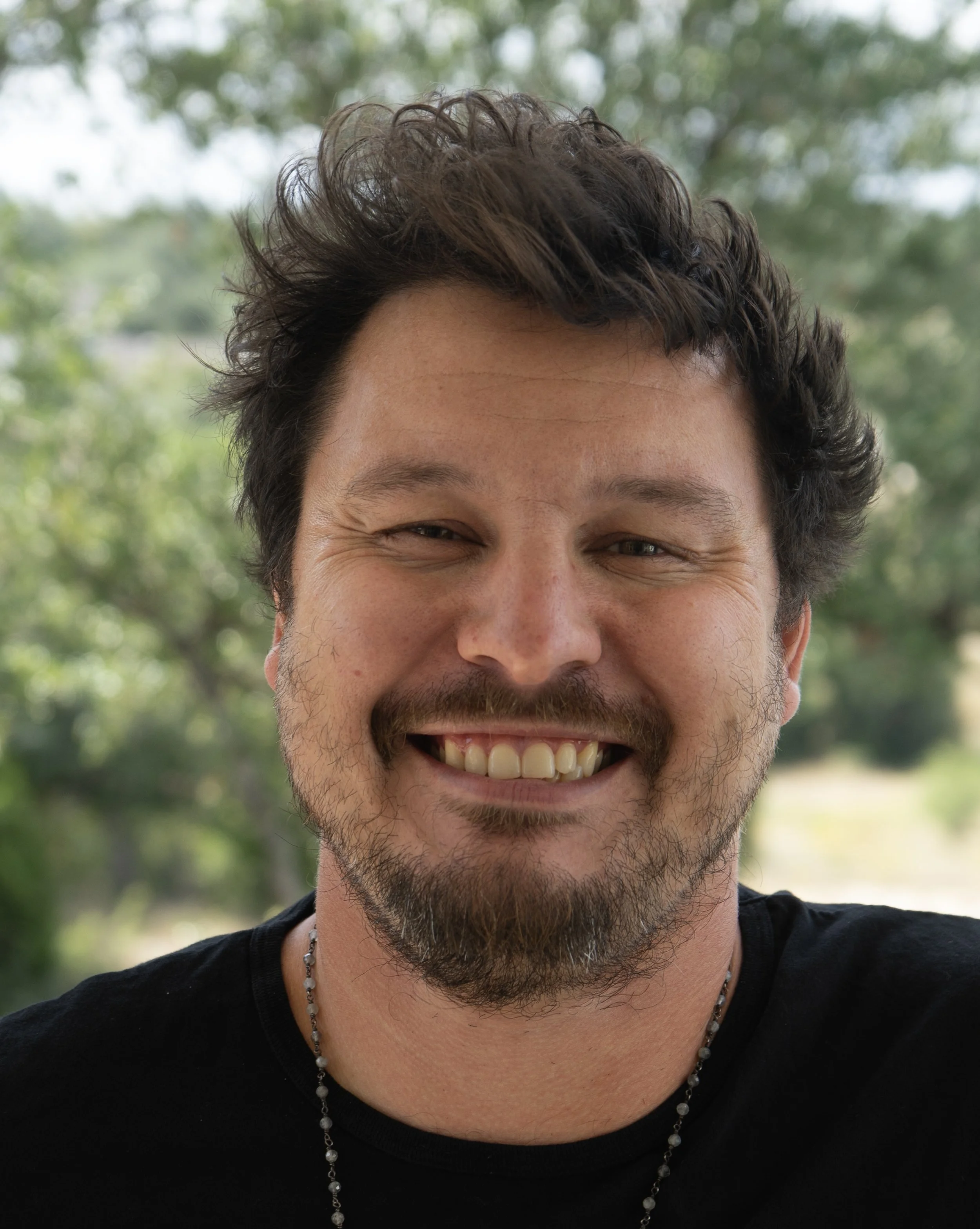Smiling man with dark, curly hair and a beard, outdoors with trees in the background.