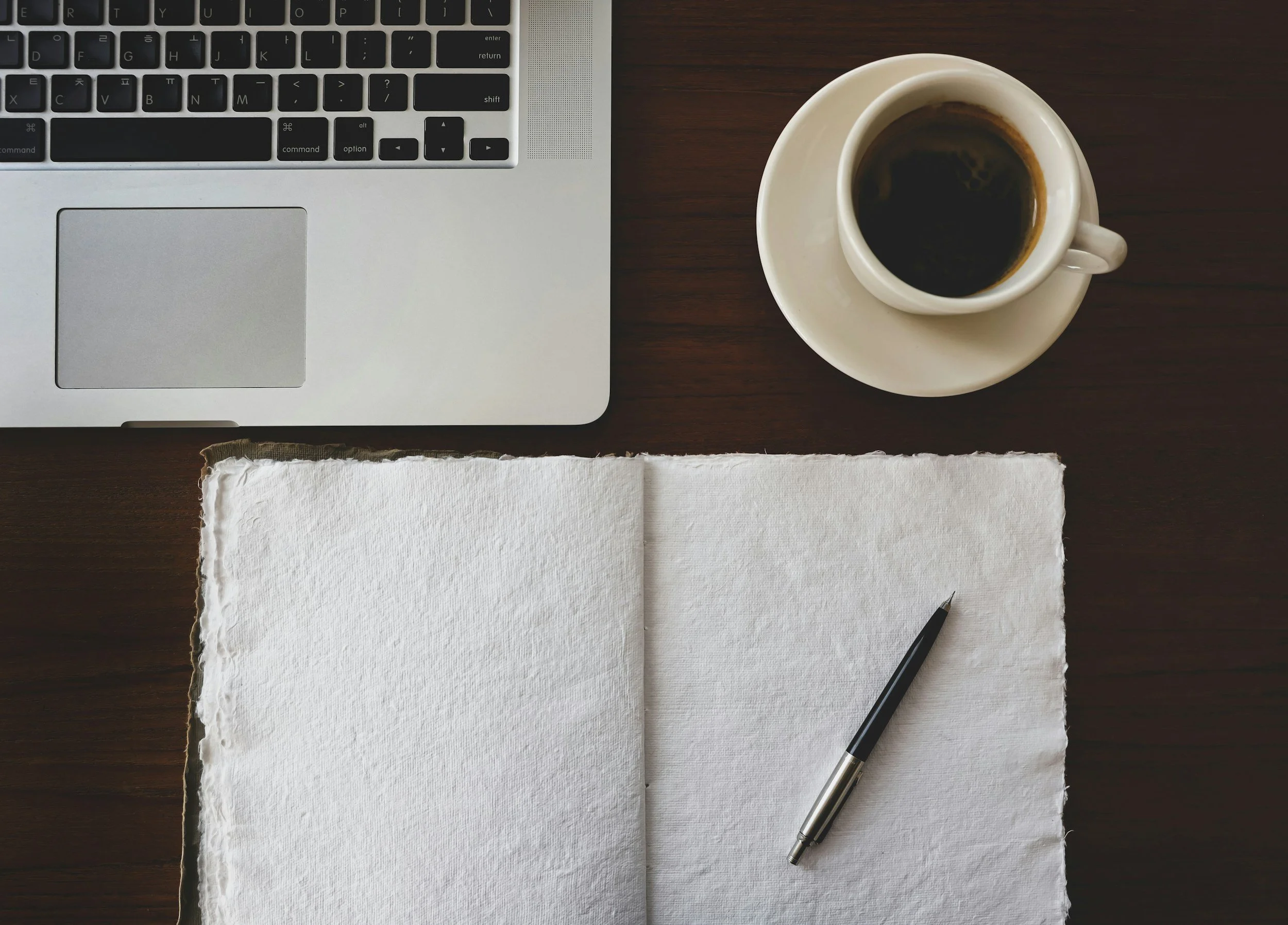 Open laptop on a wooden desk with a blank journal and a pen beside a cup of coffee.