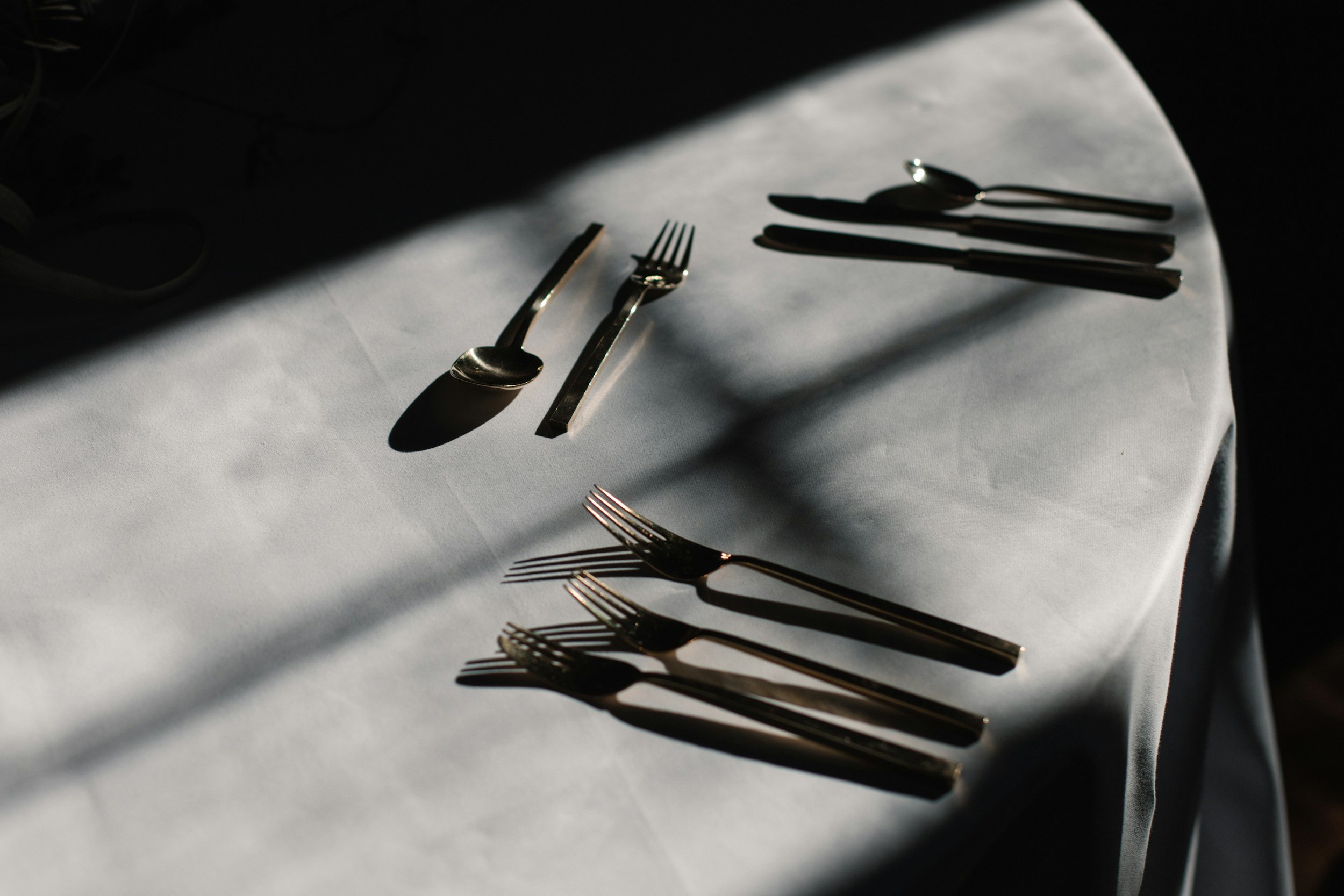 Table set with silverware on a white tablecloth in sunlight, featuring forks, knives, and spoons.