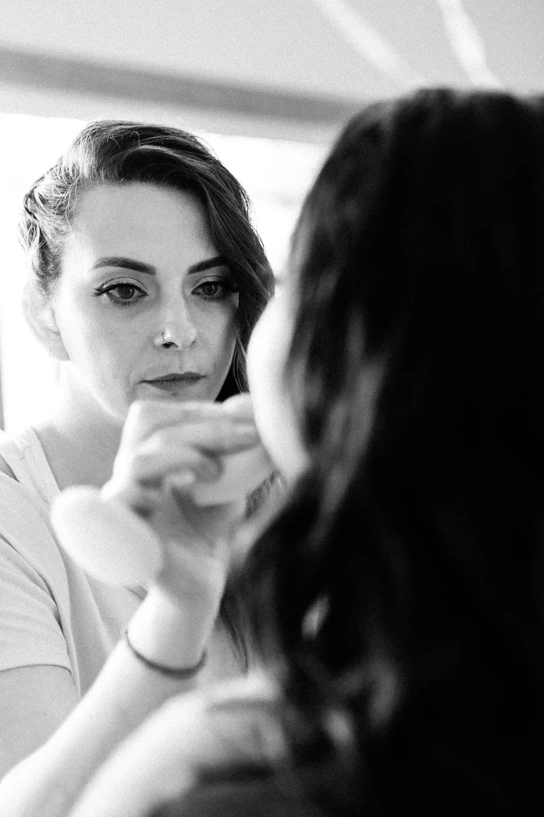 A makeup artist applies makeup to a woman's face while looking into a mirror, with a focus on her concentrated expression.