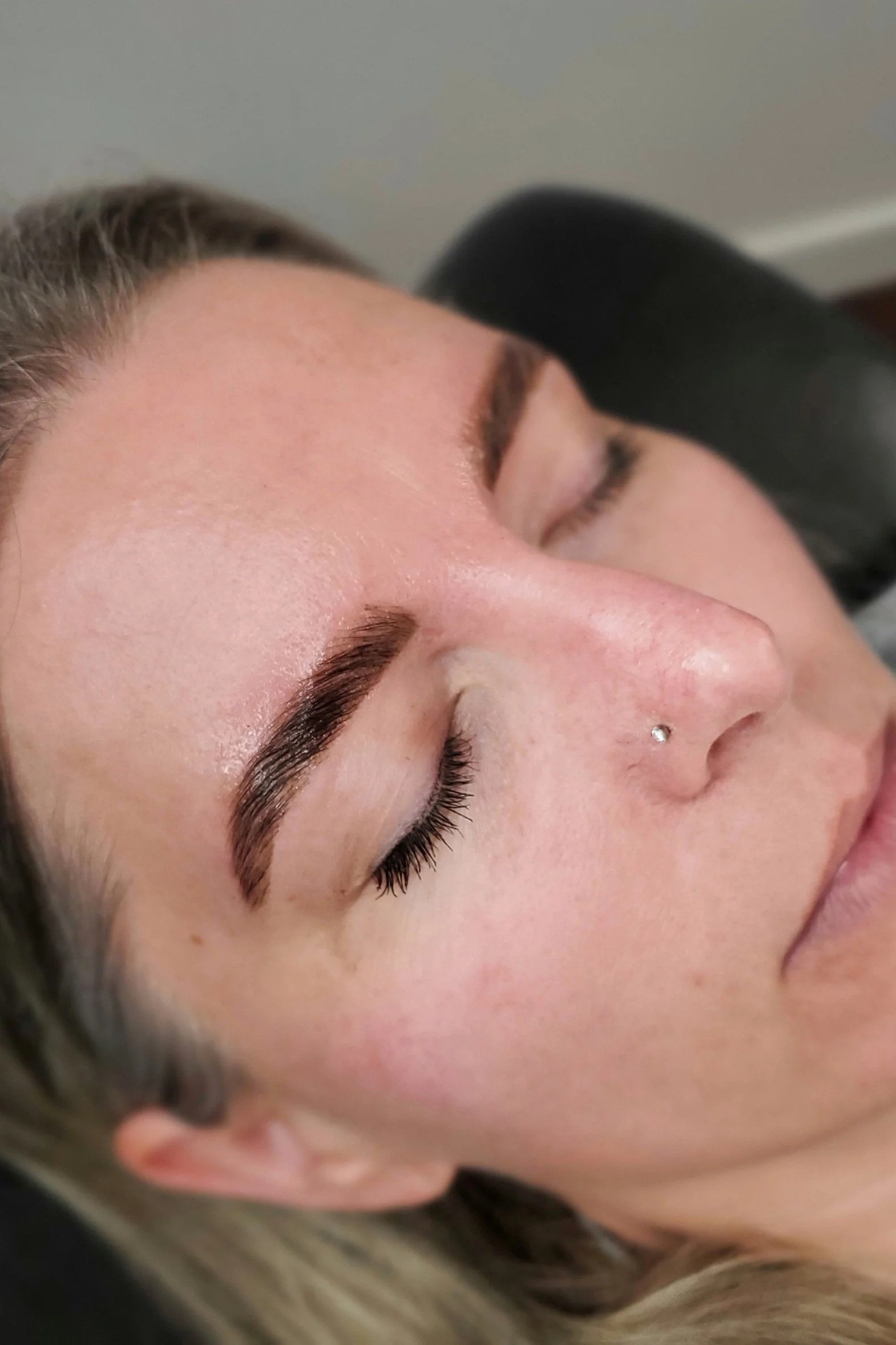 Close-up of a woman with well-groomed eyebrows and a nose piercing, lying down with her eyes closed.
