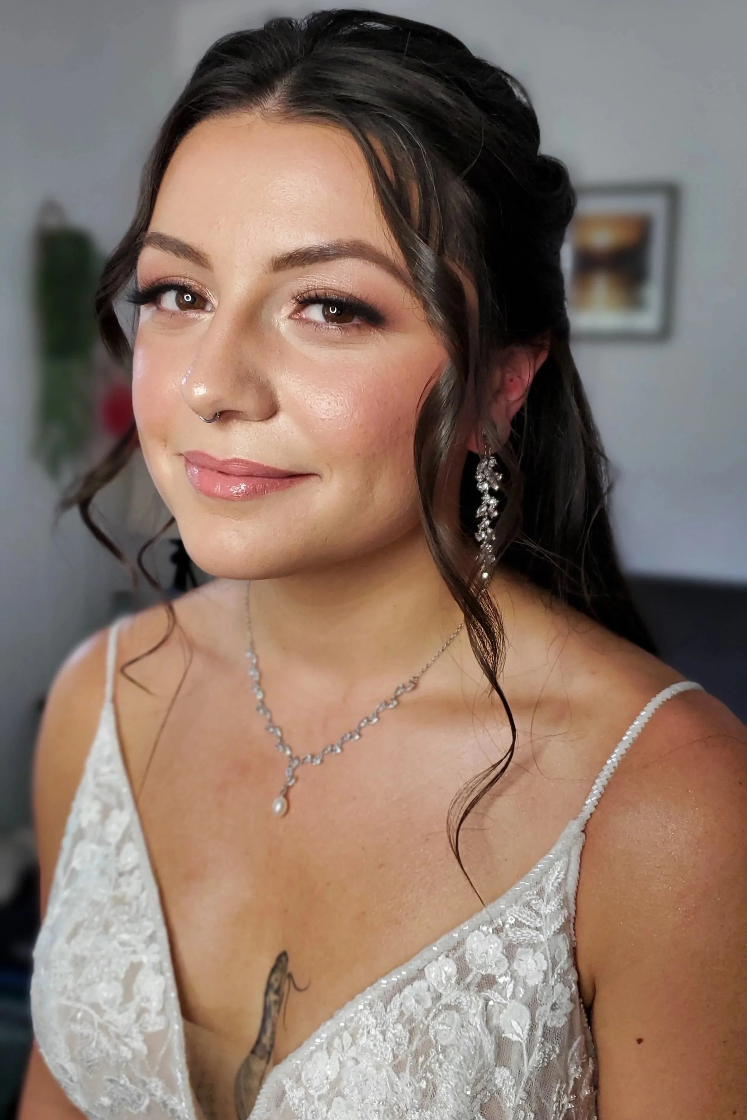 A young woman with styled dark hair and light makeup, wearing a white lace dress, jewelry, and earrings, smiling softly.