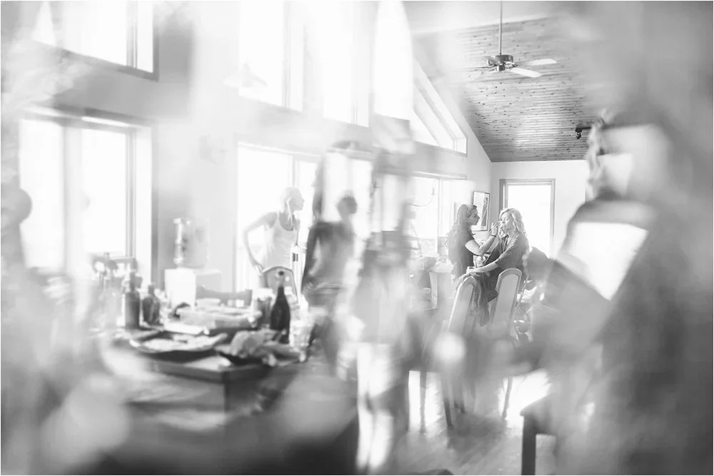 People in a bright, airy room with large windows are preparing for an event, with some people sitting and others standing and chatting.