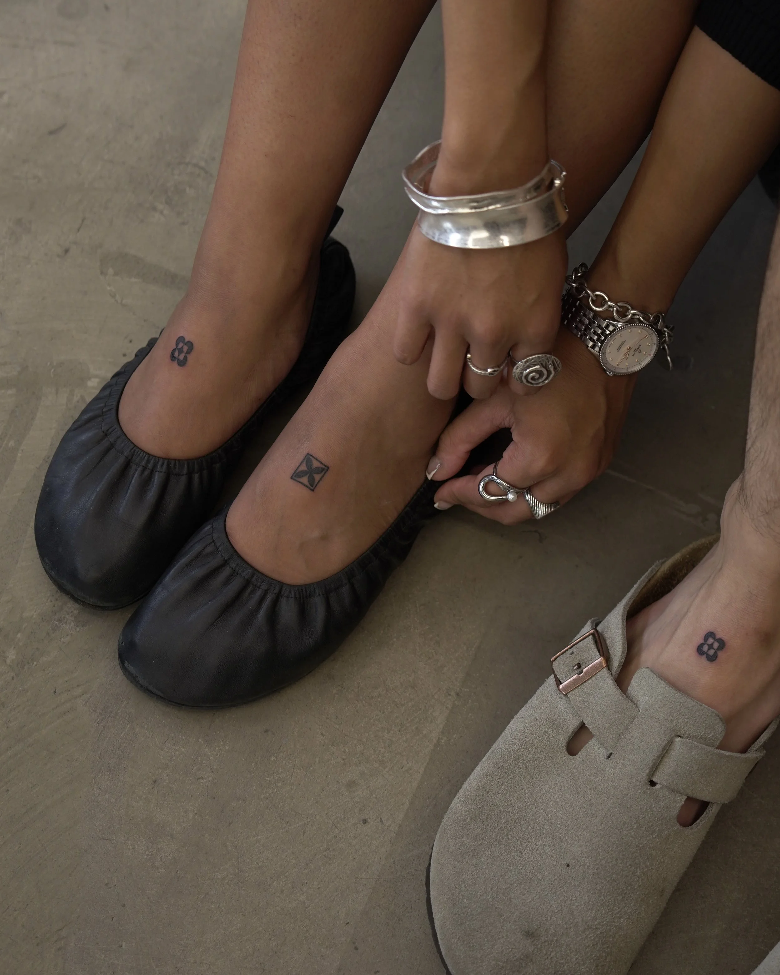 Footwear and tattoos on ankles of two people; one wearing black ballet flats with a tattoo of a four-leaf clover, and the other wearing a beige shoe with a tattoo of a four-leaf clover; accessories include rings, a silver bracelet, a watch, and a cha
