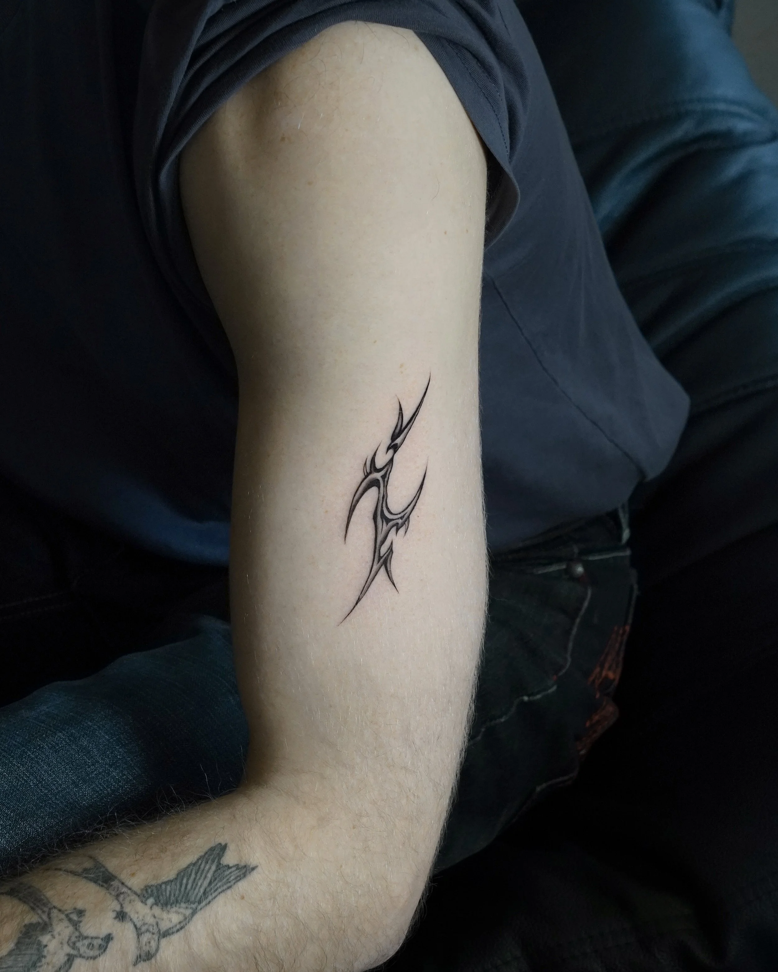 Tattoo of abstract tribal design on inner forearm of person wearing dark T-shirt, dark jeans, and with a bird tattoo on wrist.