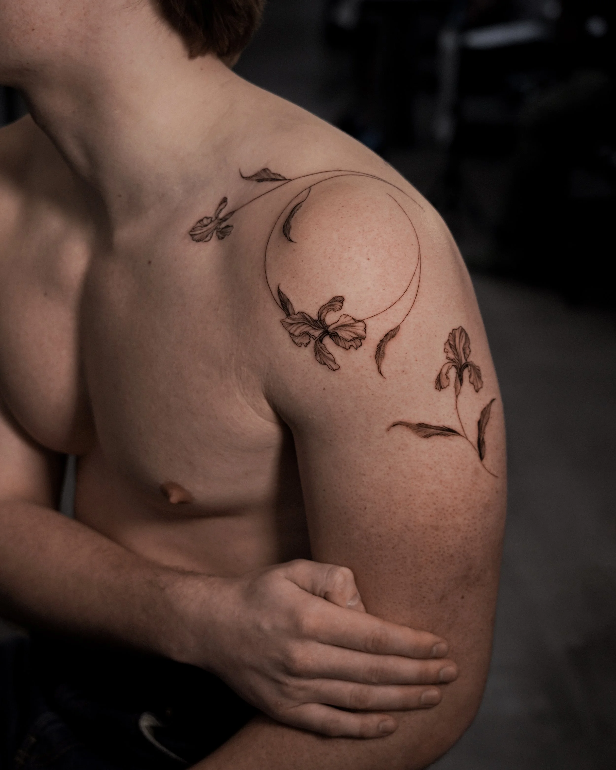 A person's upper arm and shoulder with a tattoo of a crescent moon, butterflies, and leaves, in a minimalistic line art style.