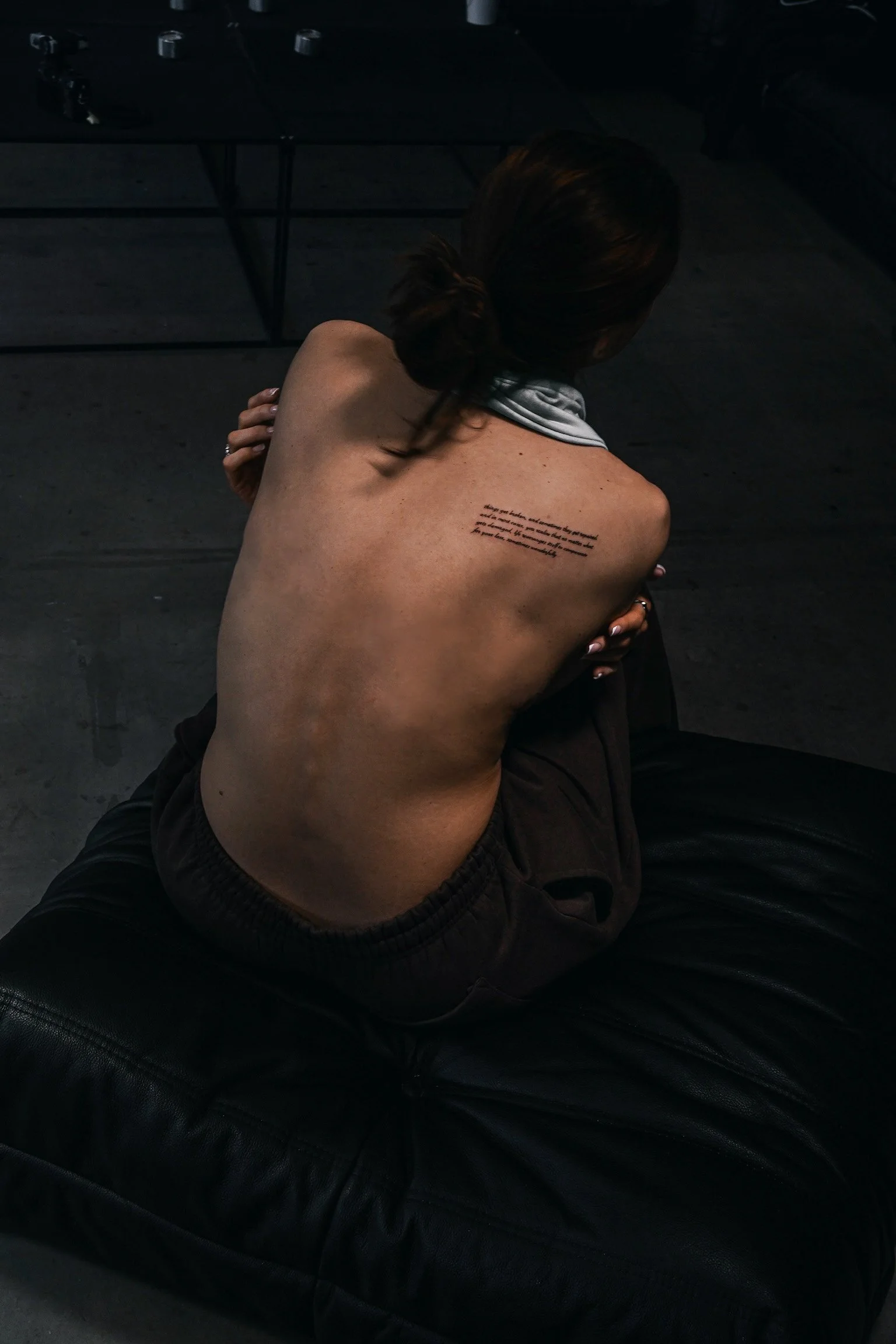 A woman with dark hair sitting on a black leather chair, with her back turned to the camera, showcasing a tattoo on her upper back.
