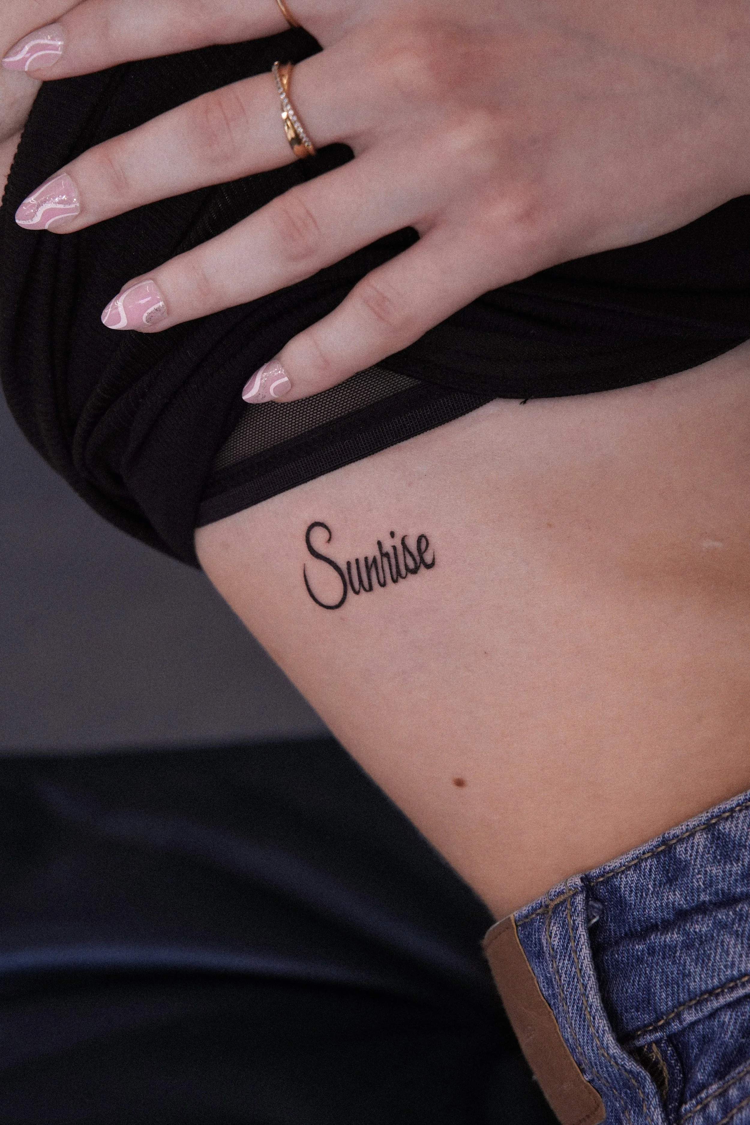 A person shows a tattoo on their ribcage that reads "Sunrise" in black cursive font, with fingers resting on their black shirt.