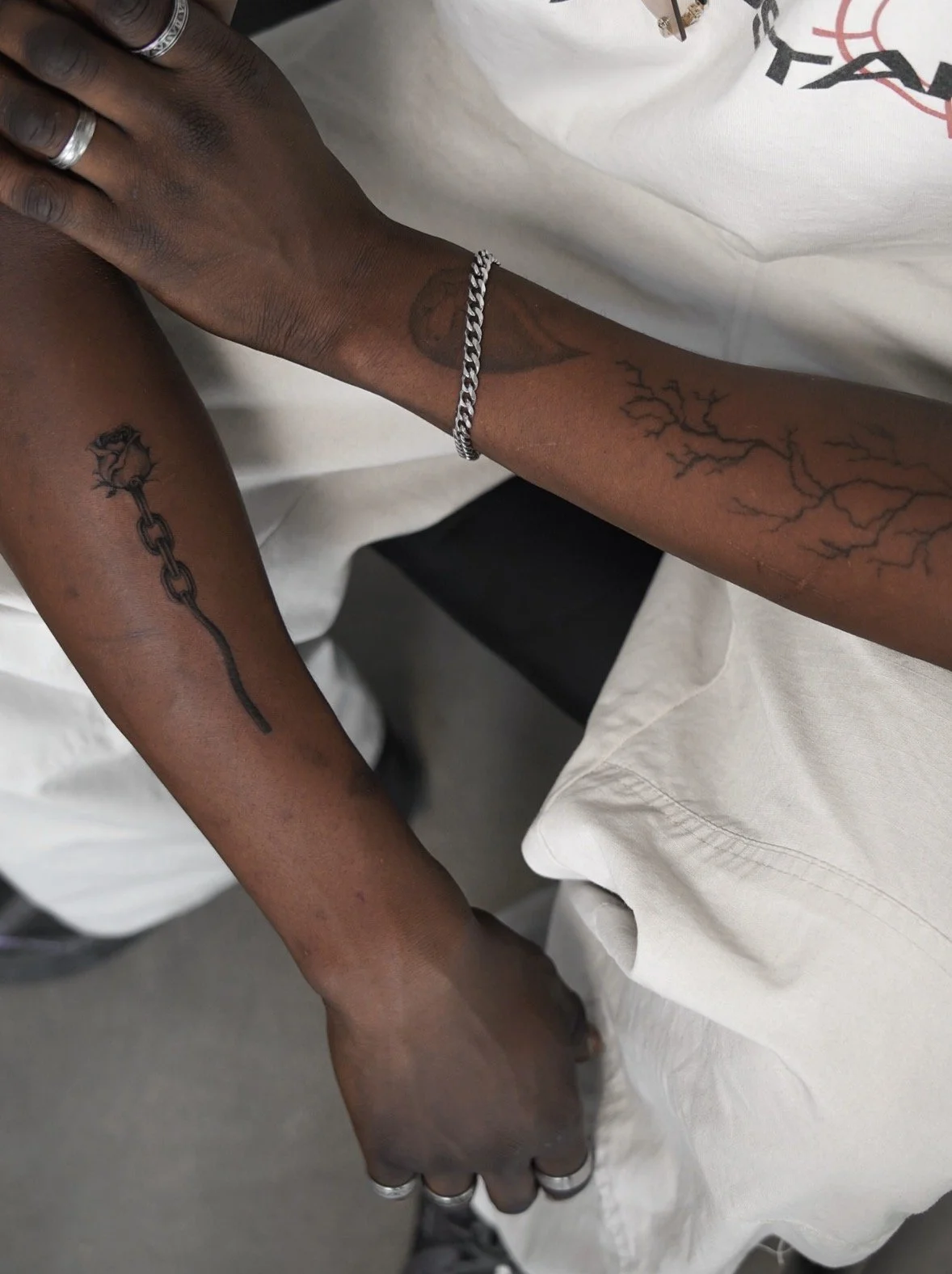 Close-up of a person's arm with tattoos, one of a branch, one of a broken chain, and one of a tree. The person wears a silver bracelet and rings and has other jewelry. The person is wearing a white T-shirt and beige pants.