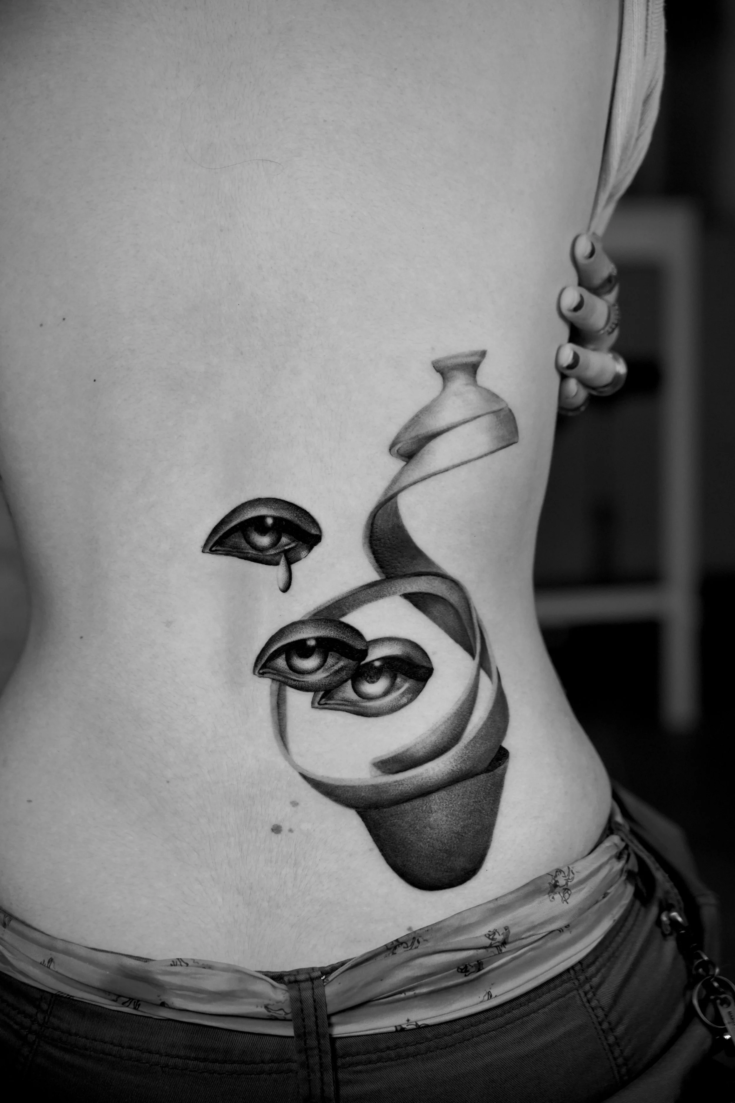 Black and white tattoo of three human eyes with teardrops on a person's side torso, with a spiral abstract design above the eyes.