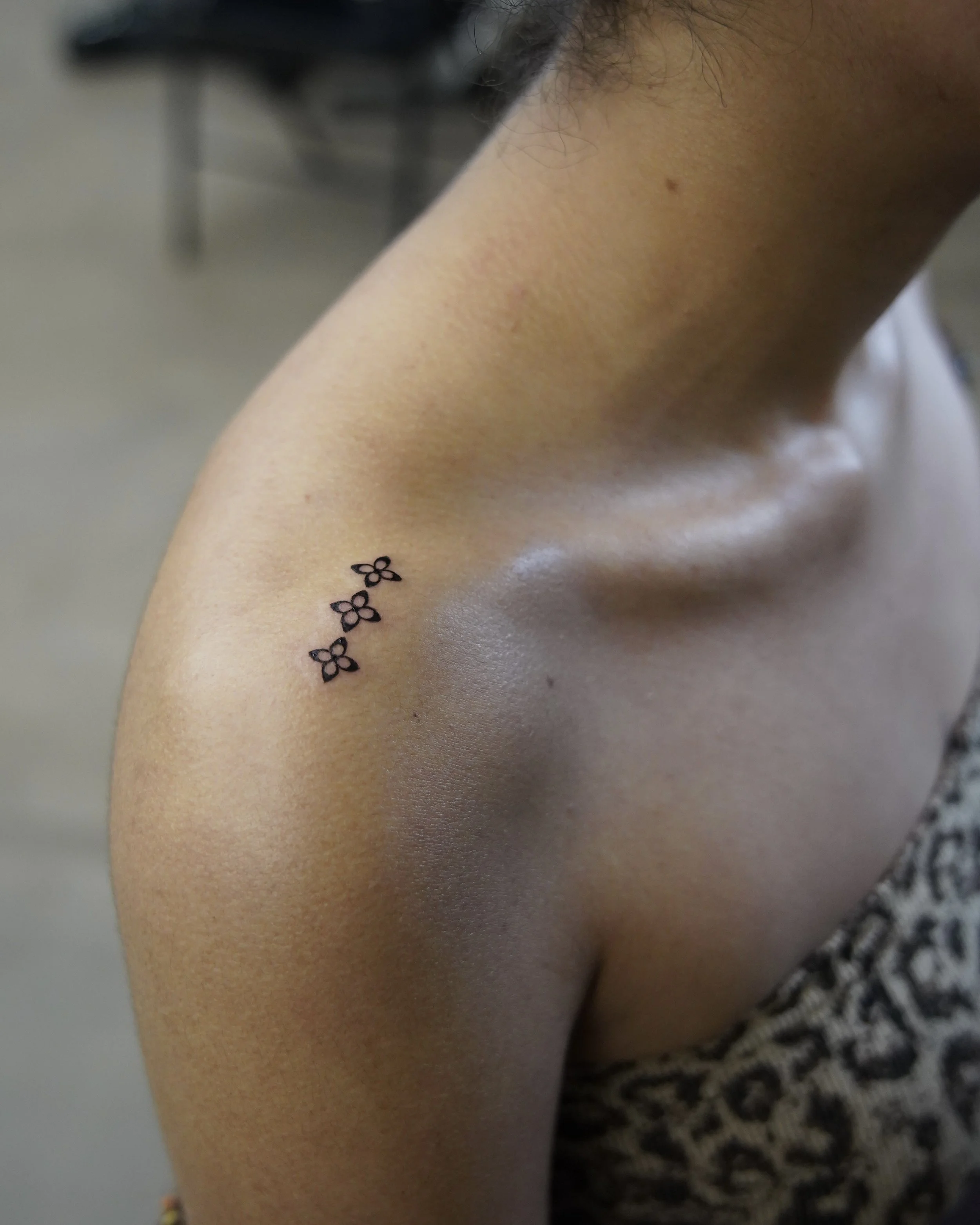 A close-up of a person’s shoulder and neck with a small tattoo of three flowers on the upper arm near the shoulder.