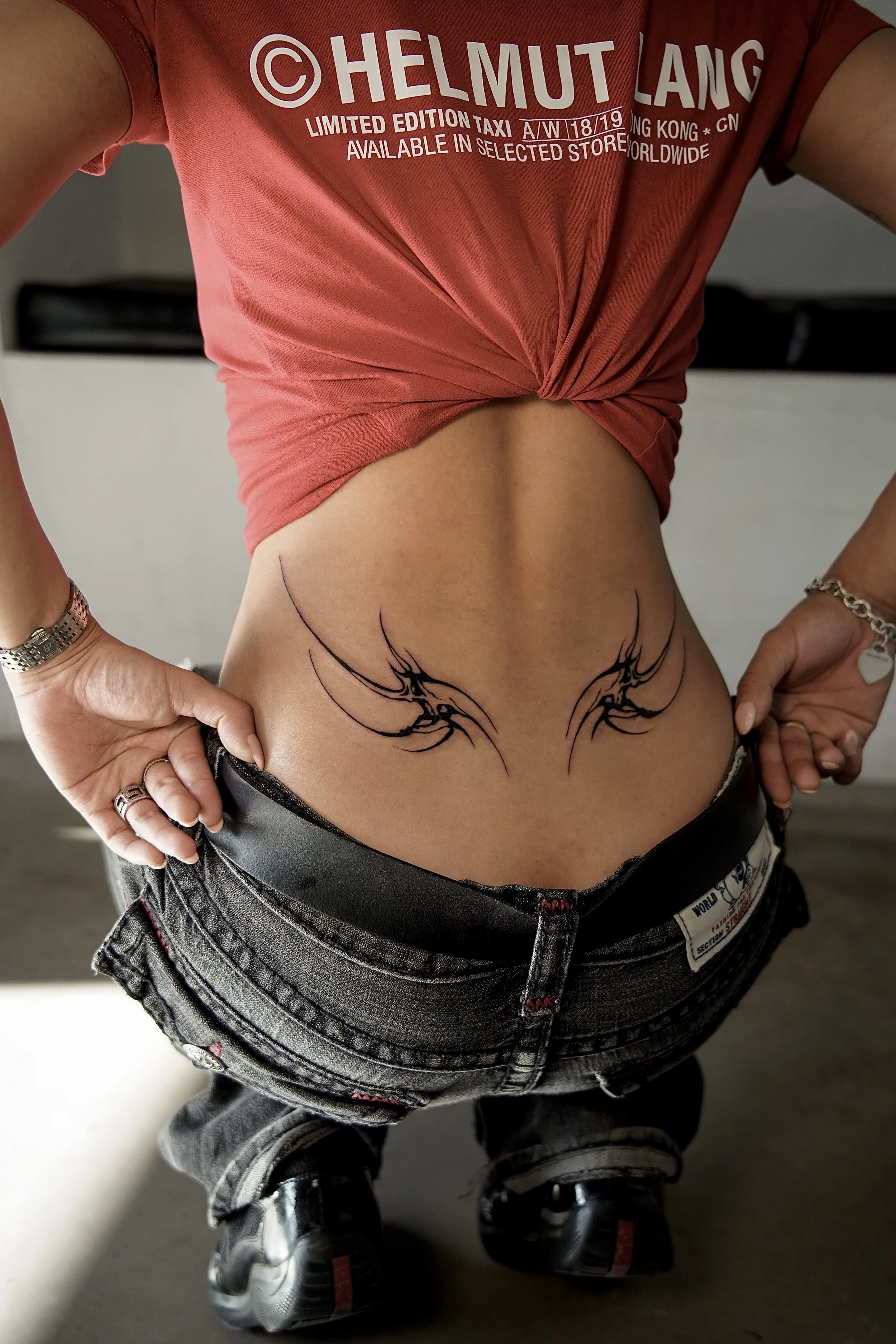 A person standing with hands on hips, wearing a knotted pink T-shirt revealing a tattoo of two black abstract wings on their lower abdomen, black jeans, and black sneakers, with living room background.