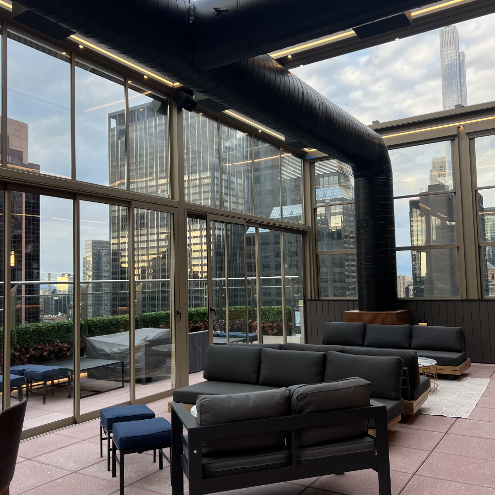 Interior of a modern rooftop lounge with black leather sofas, small blue cushioned stools, large windows showing a city skyline of tall buildings, and a large black duct running along the ceiling.
