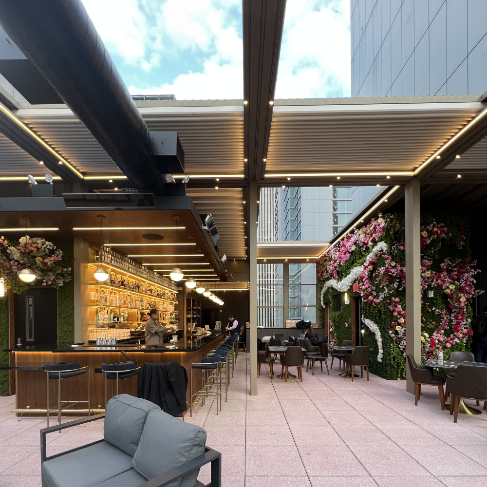 Indoor rooftop bar with a pink tiled floor, floral wall decor on the right, modern furniture, and a bar with wine bottles on shelves. Large windows and a partially open ceiling with city buildings and blue sky visible.