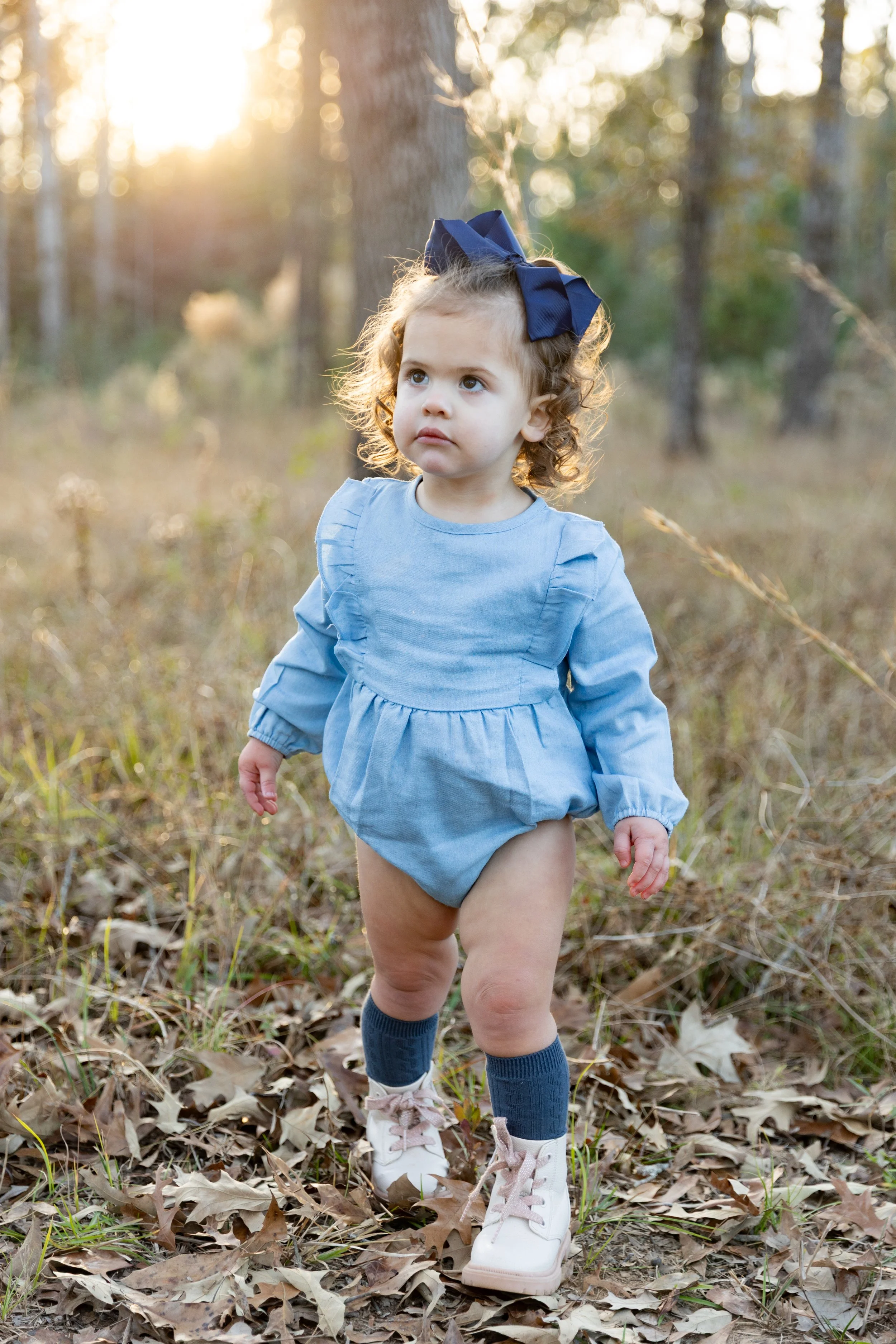 A young girl with curly hair, blue dress, blue socks, and pink shoes walking through a wooded area with fallen autumn leaves, during sunset.