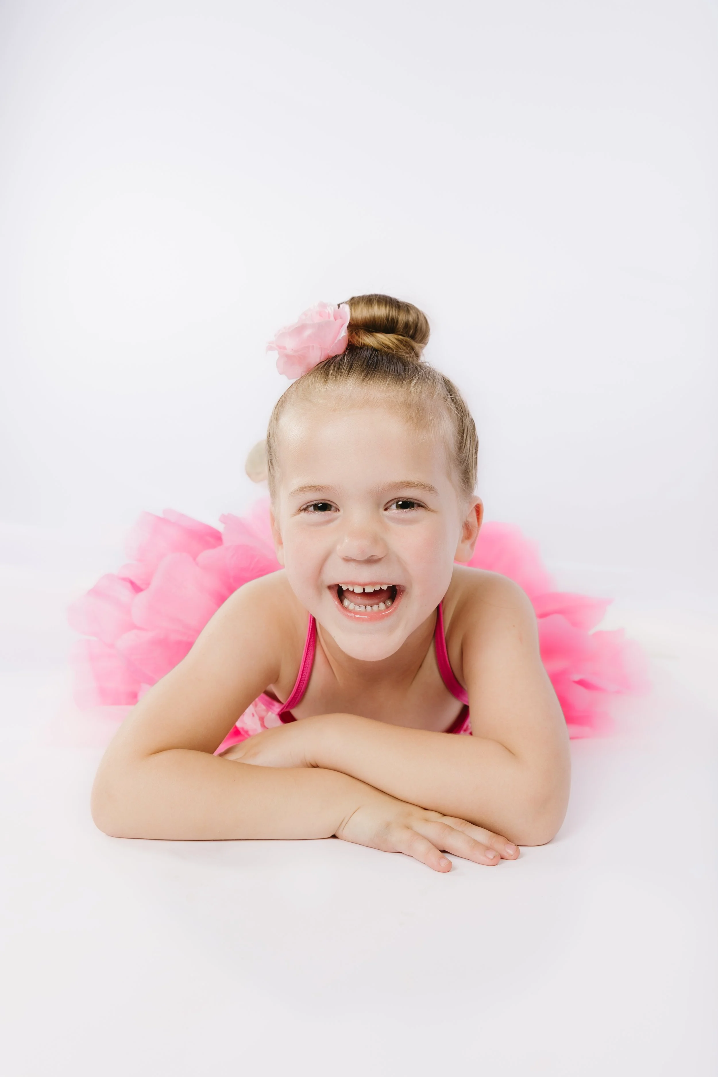 A young girl with a smile, lying on her stomach on a white background, decorated with a pink flower hair accessory and pink tutu, smiling at the camera.