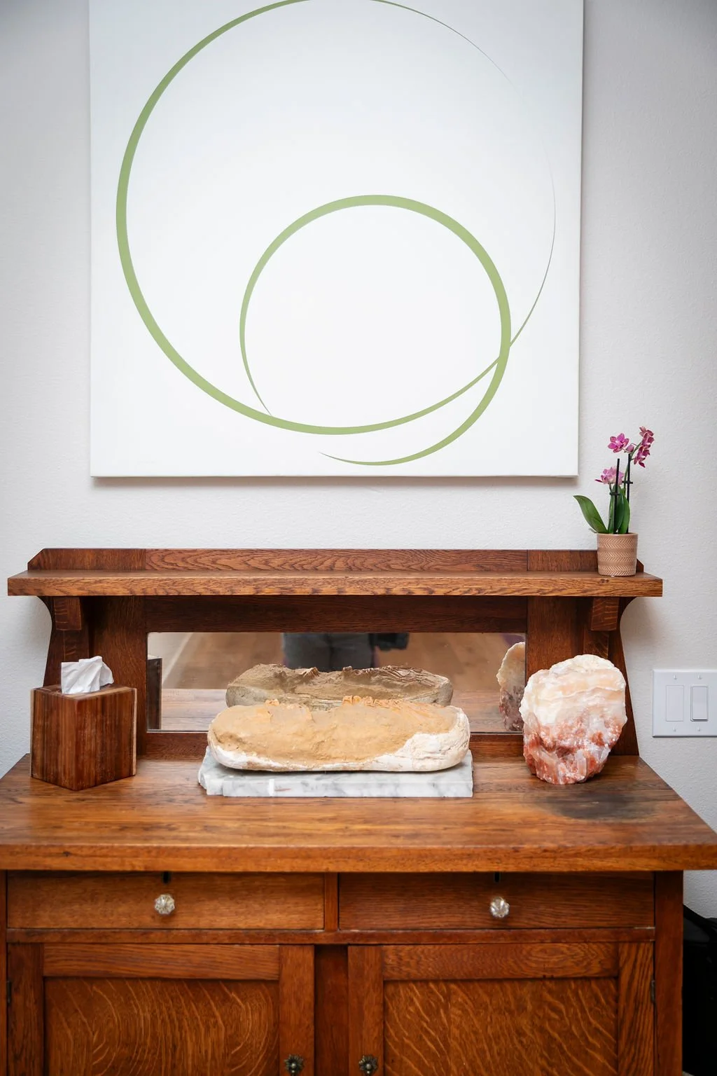 A wooden sideboard with mineral stones and a mirror. Above, a white wall art with green circles, and a small potted pink orchid plant.