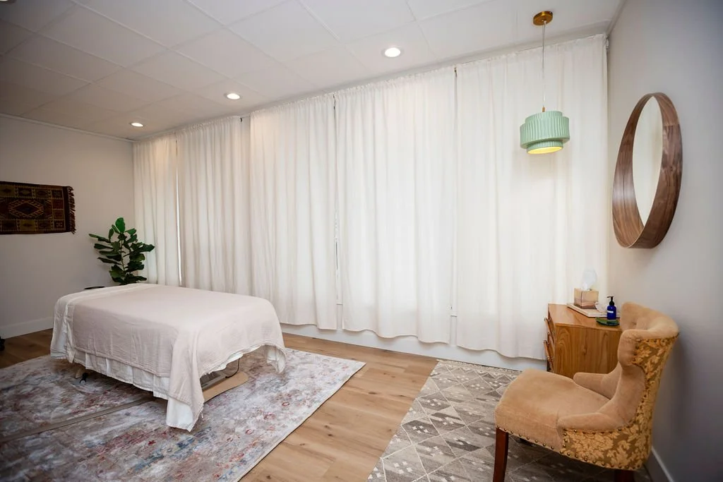 A bedroom with a bed covered in a white quilt, a vintage armchair, a wooden side table with a tissue box and a blue bottle, a round mirror on the wall, and white curtains covering large windows.