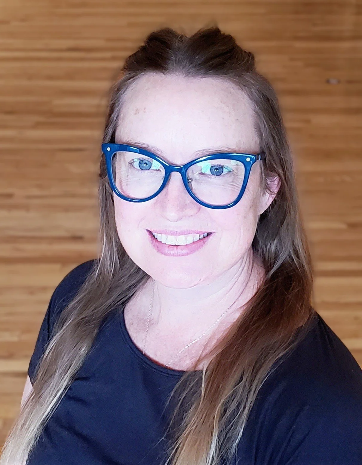 A woman with long brown hair, blue glasses, and a black shirt, smiling for a selfie in front of a wooden background.