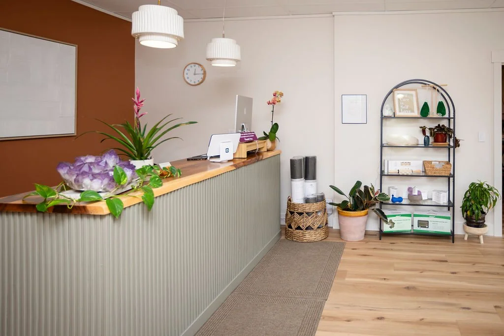 Reception area with a wooden front desk, potted plants, shelves with decorative items, and a wall clock.