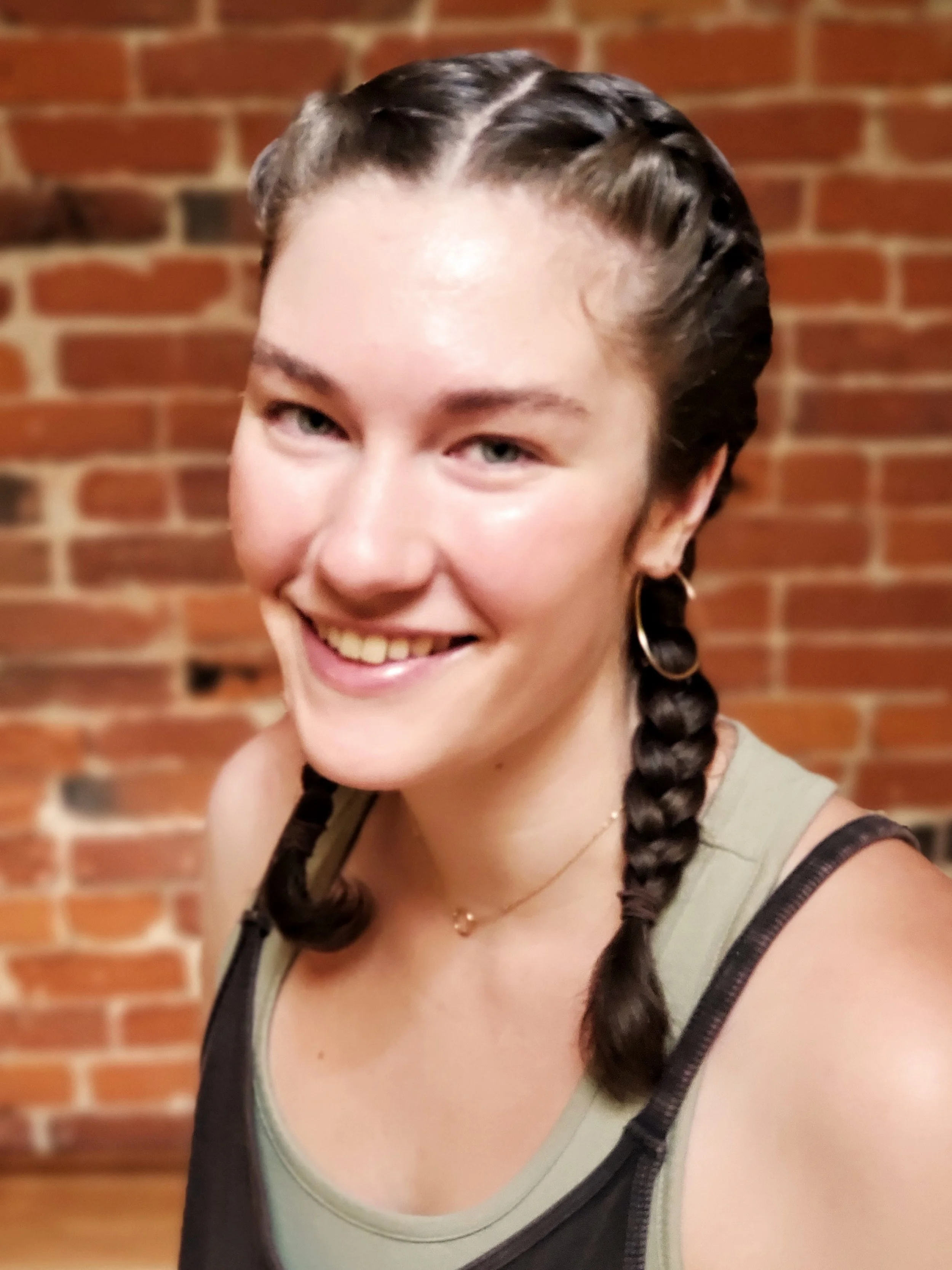 Young woman smiling with braided hair, wearing hoop earrings and a necklace, standing in front of a brick wall.