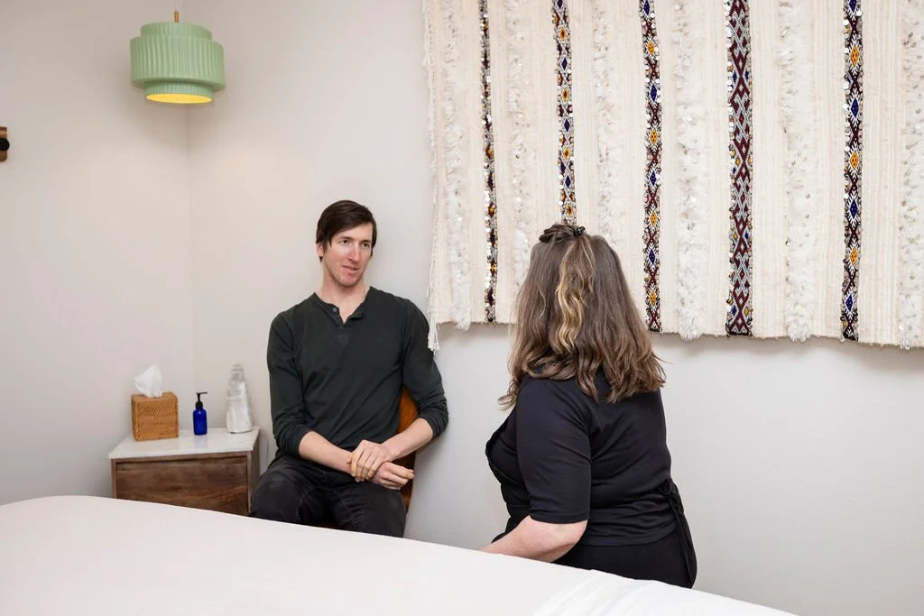 A man and woman sit in a room, engaging in conversation. The man has dark hair, wears a black shirt, and is sitting on a chair. The woman has shoulder-length hair, wears a black shirt, and is sitting on a bed or similar surface. A small side table wi