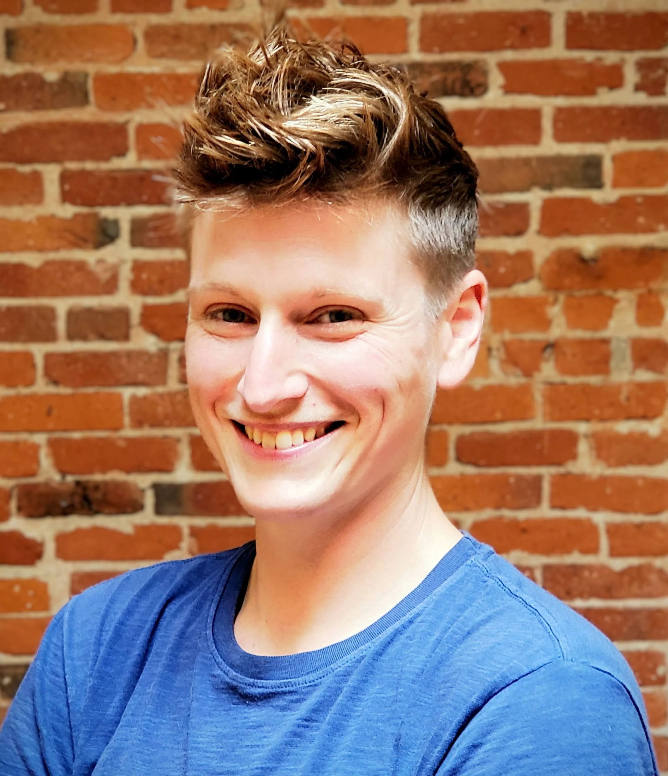 A person with short, styled brown hair smiling against a brick wall, wearing a blue shirt.
