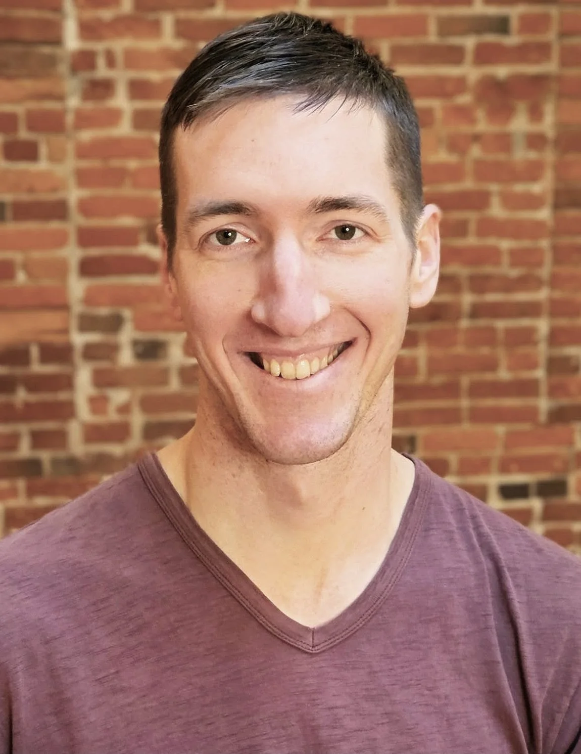 A man with short dark hair and a friendly smile, wearing a maroon V-neck shirt, standing in front of a brick wall.