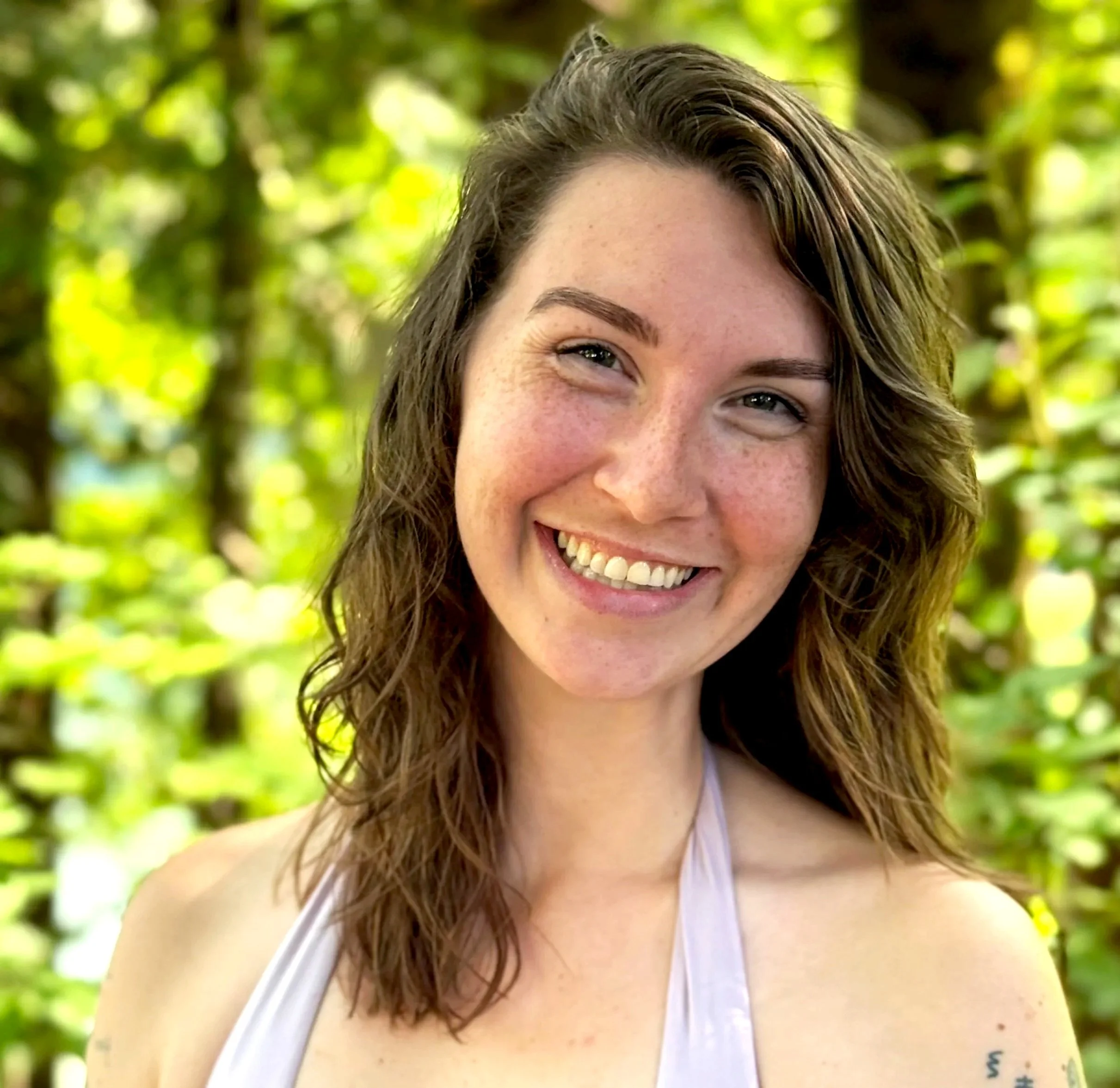 Young woman with short brown hair smiling, wearing a bright yellow sleeveless top, a colorful headscarf, and earrings, standing against a brick wall.