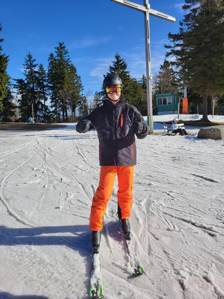 Person in ski gear standing on skis on a snowy slope, with a tall cross in the background and trees surrounding the area.