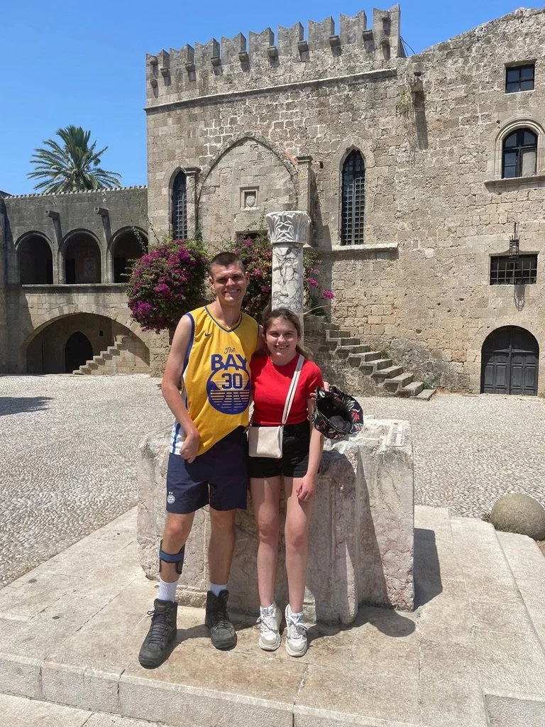 Two people standing in front of an ancient stone building with arched windows, wearing casual summer clothes and smiling, with flowers and palm trees in the background.