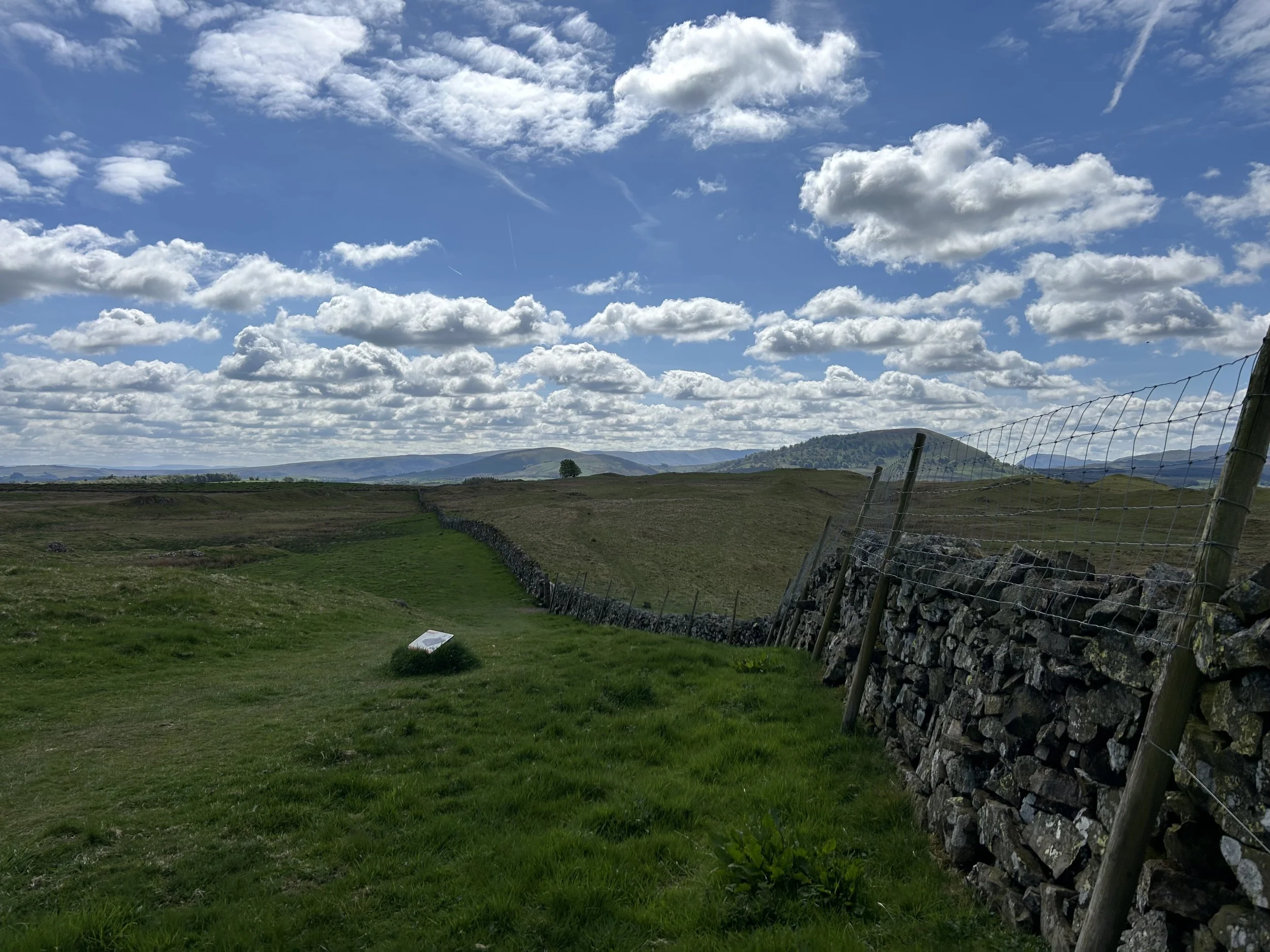 A view of a dry stone wall cutting through the middle of hills and fields. Above is a clear blue sky with some clouds in the distance.
