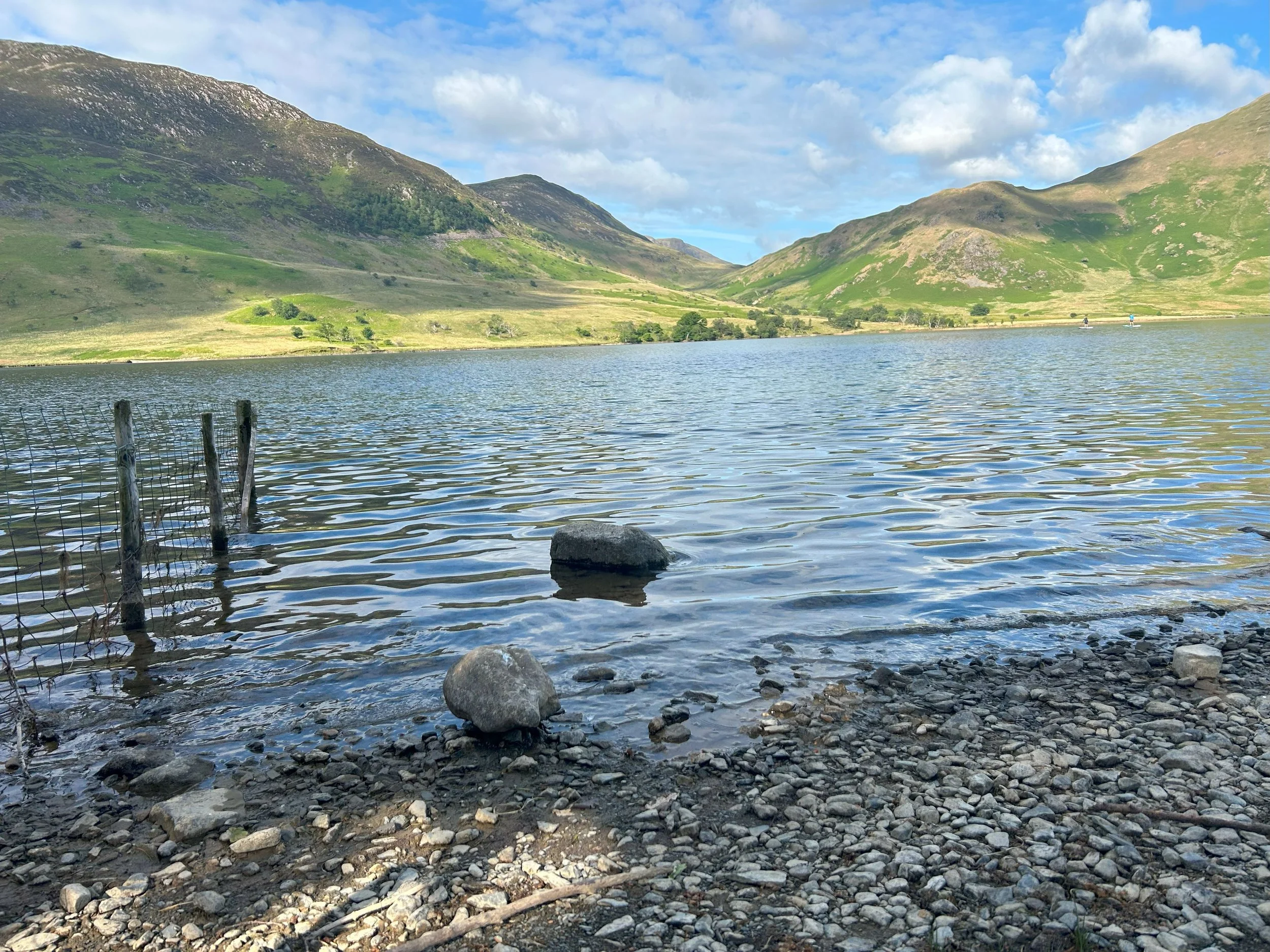 A small part of the stony beach in the foreground, leading to lake crummock and rolling mountain views in the distance. There are two paddle boarders in the far distance on the lake.