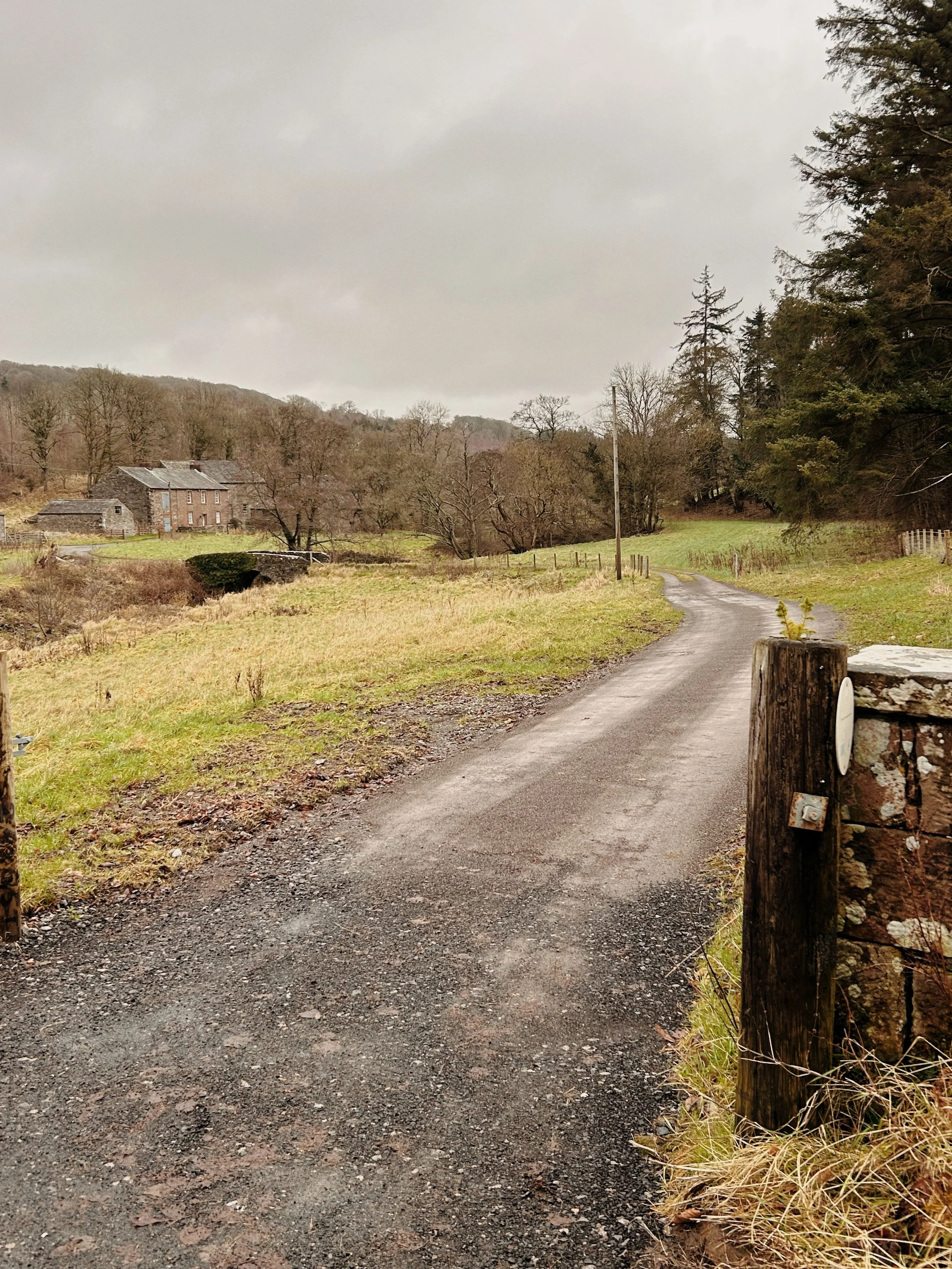 A farm track flanked by fields, leading over a small stone bridge to some creepy farm buildings. There are trees and a bleak, grey sky in the background