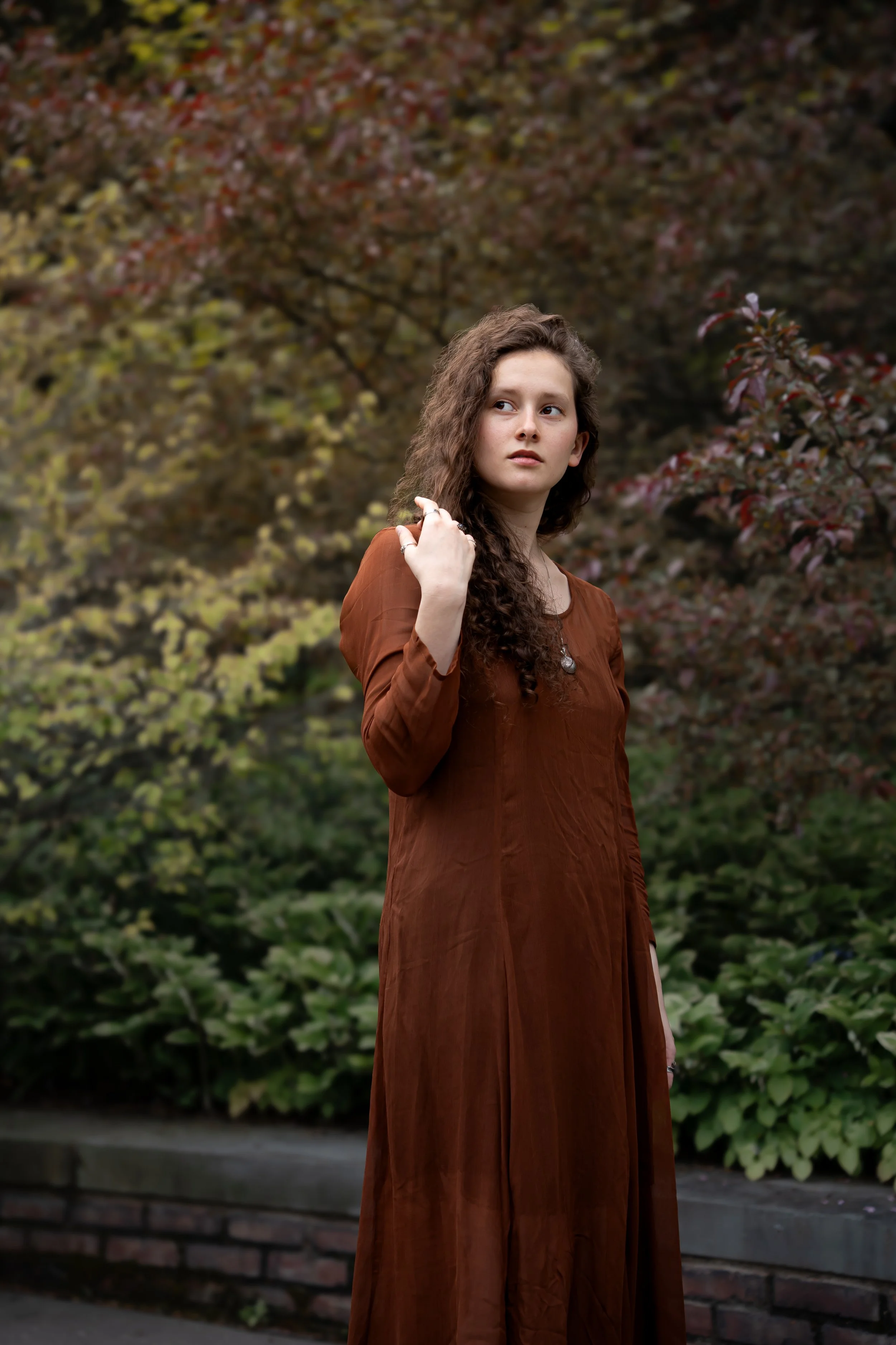 A young woman with long, curly brown hair standing outdoors in a park-like area during autumn. She is wearing a long brown dress and gazing to her right with a neutral expression. The background has trees with reddish leaves and some greenery.