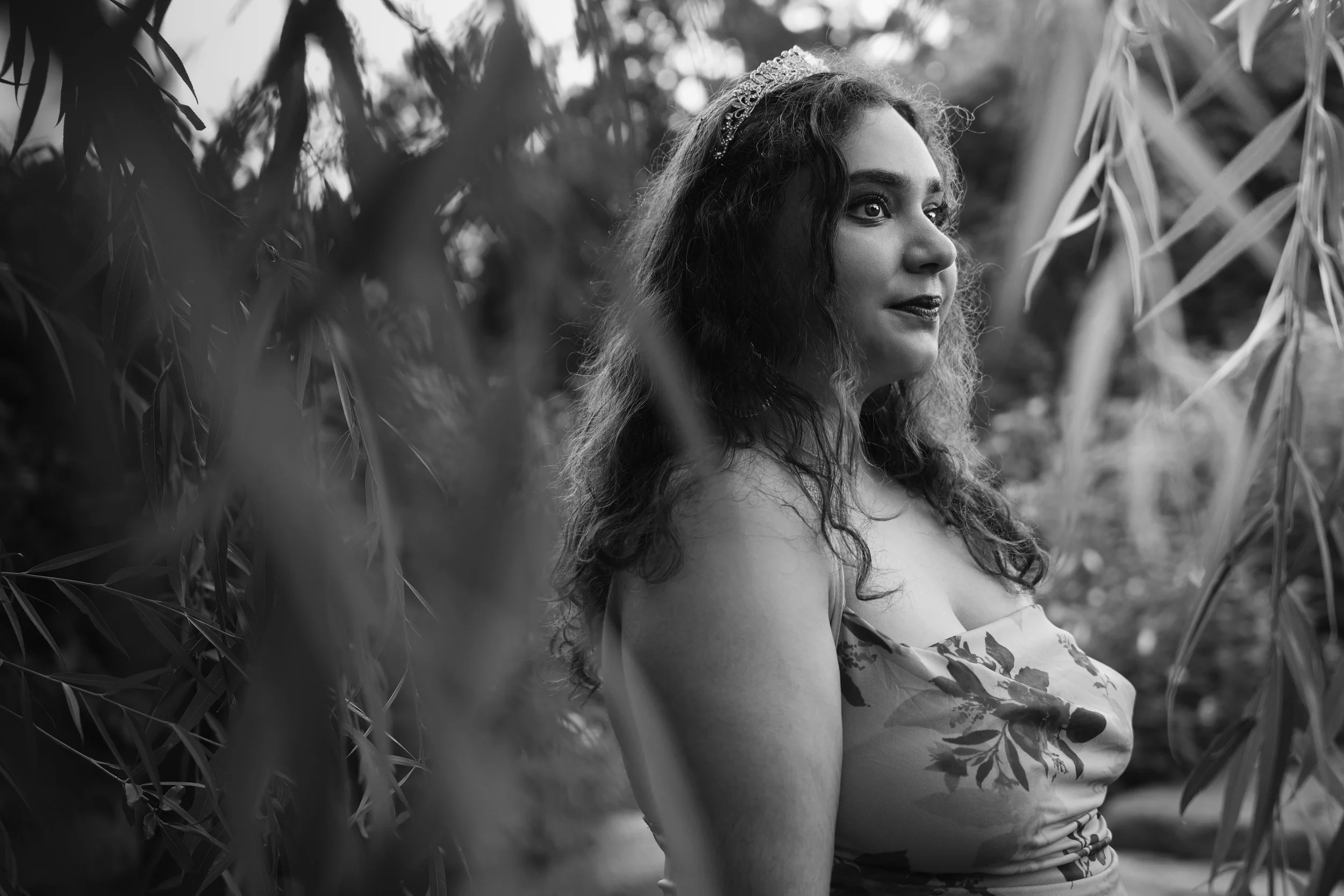 A woman with long, curly hair wearing a tiara and floral dress, sitting among tall plants in an outdoor setting, captured in black and white.