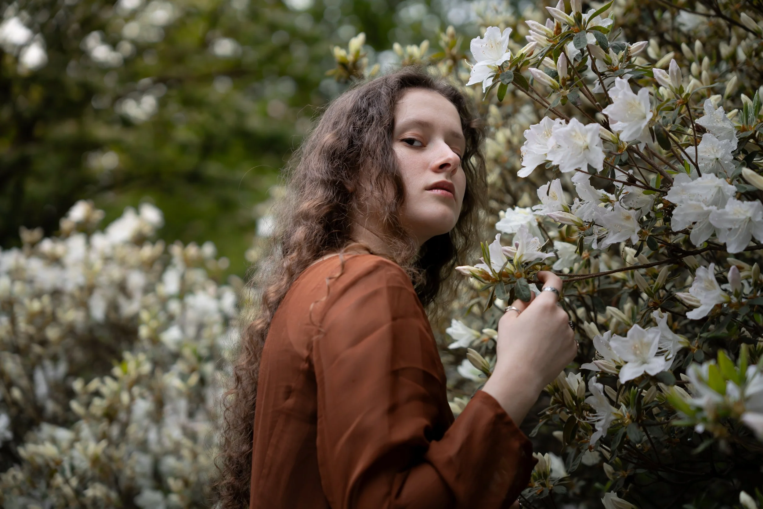 A woman with long curly hair and a brown jacket standing among white flowering bushes, looking at the camera.