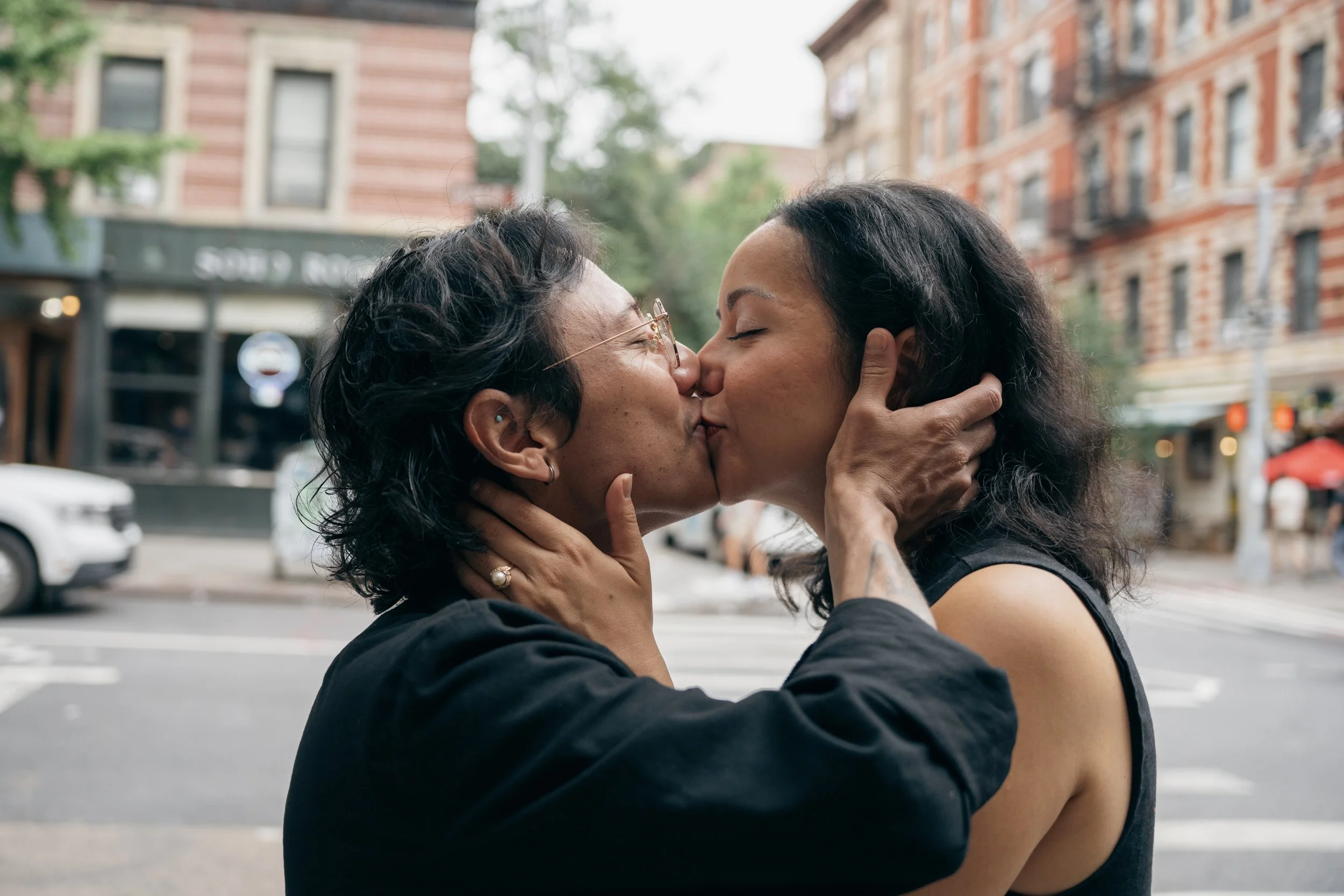 Two women share a kiss on a city street, holding each other's faces. The woman on the left has short gray hair, glasses, and earrings; the woman on the right has long dark hair. Buildings and trees are visible in the background.