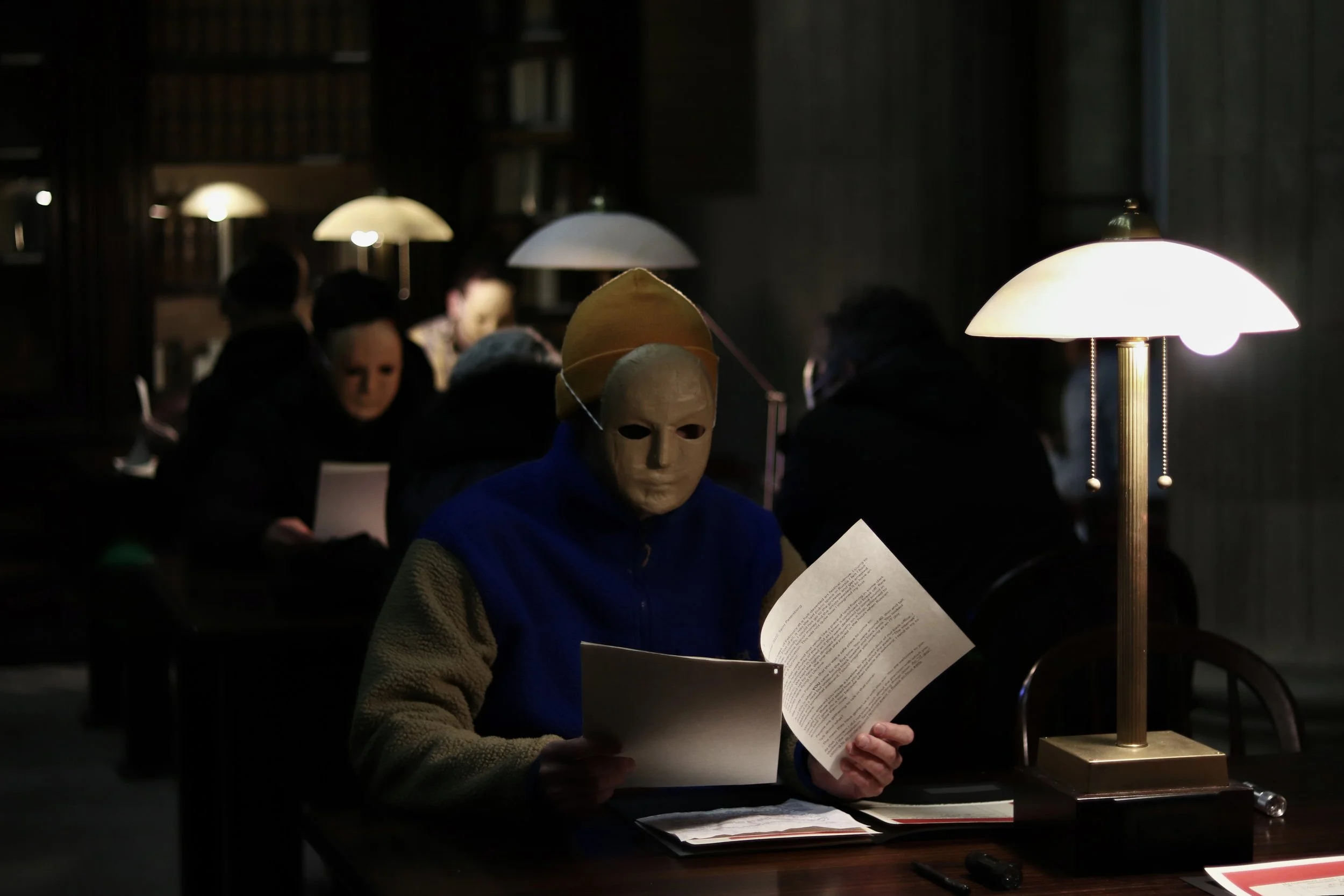 A person wearing a white mask with hollow eyes and nose, dressed in a blue shirt and beige jacket, sitting at a dimly lit table reading a document. There are multiple white lamps and other people in the background.