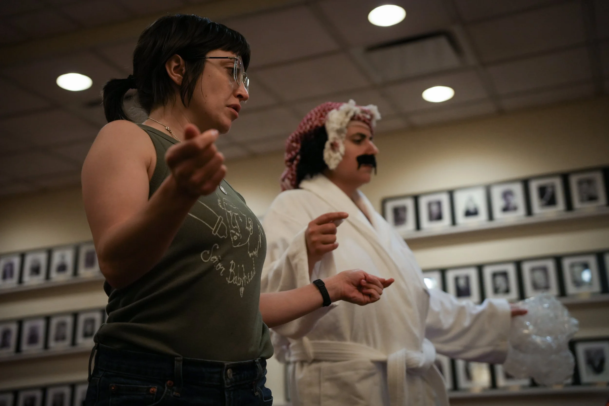 A woman wearing glasses and a sleeveless shirt with graphic print, standing with her eyes closed and hands clasped, appears to be praying or meditating. In the background, another woman dressed as a chef with a pink and white hat and white uniform al